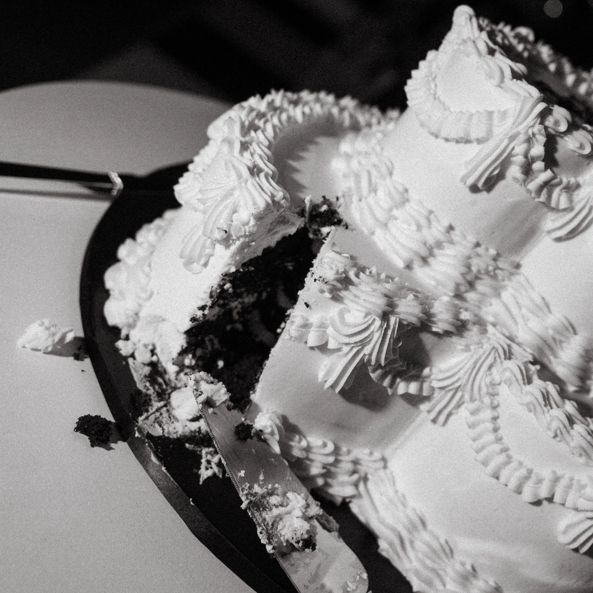 Black and white photo of a birthday cake with white frosting and decorative piping, with a slice removed revealing dark cake layers inside, placed on a white surface.