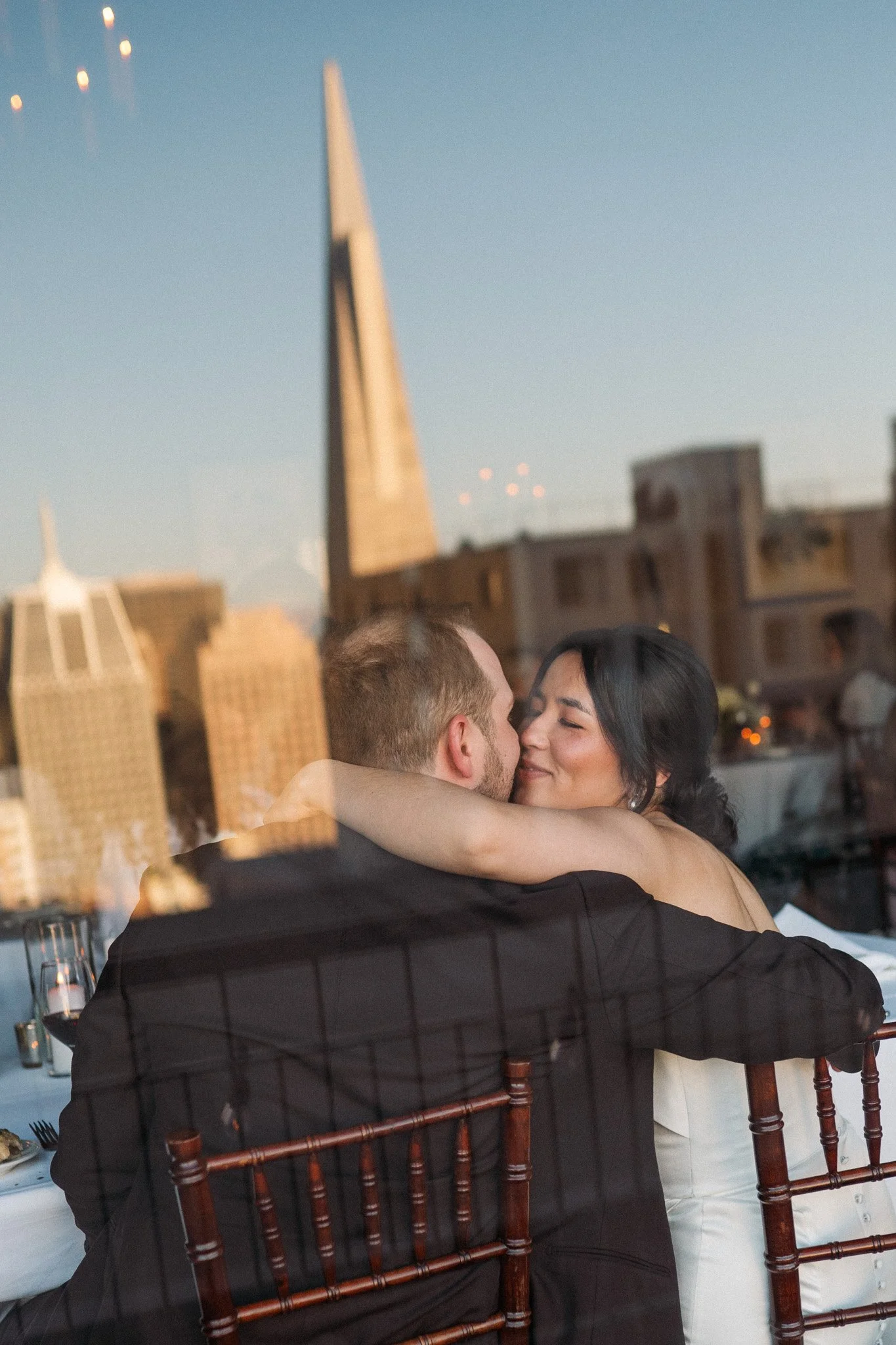 Couple embracing at a city restaurant with skyline reflections in the window.