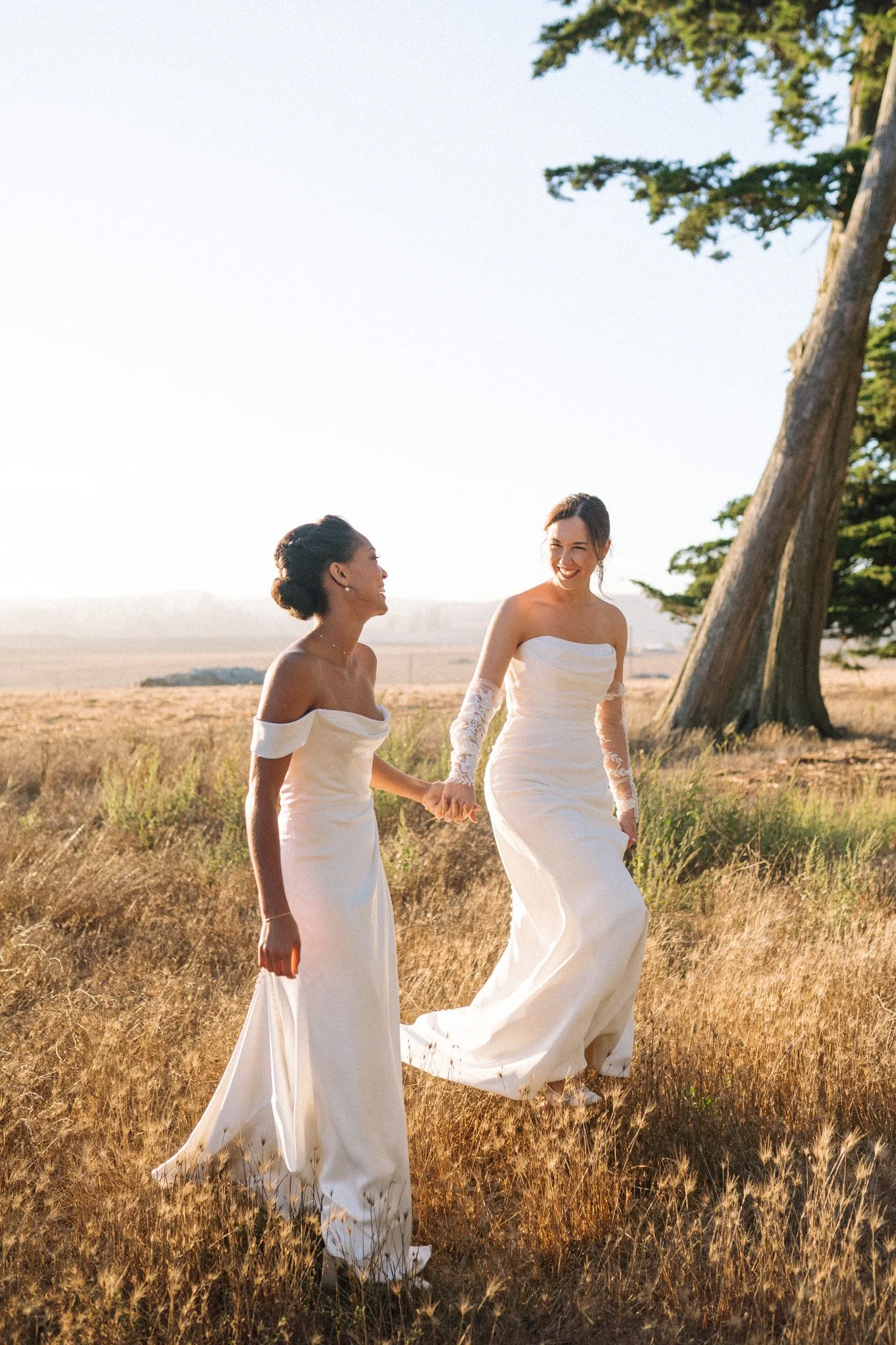Two women in wedding dresses holding hands and walking through a field at sunset.