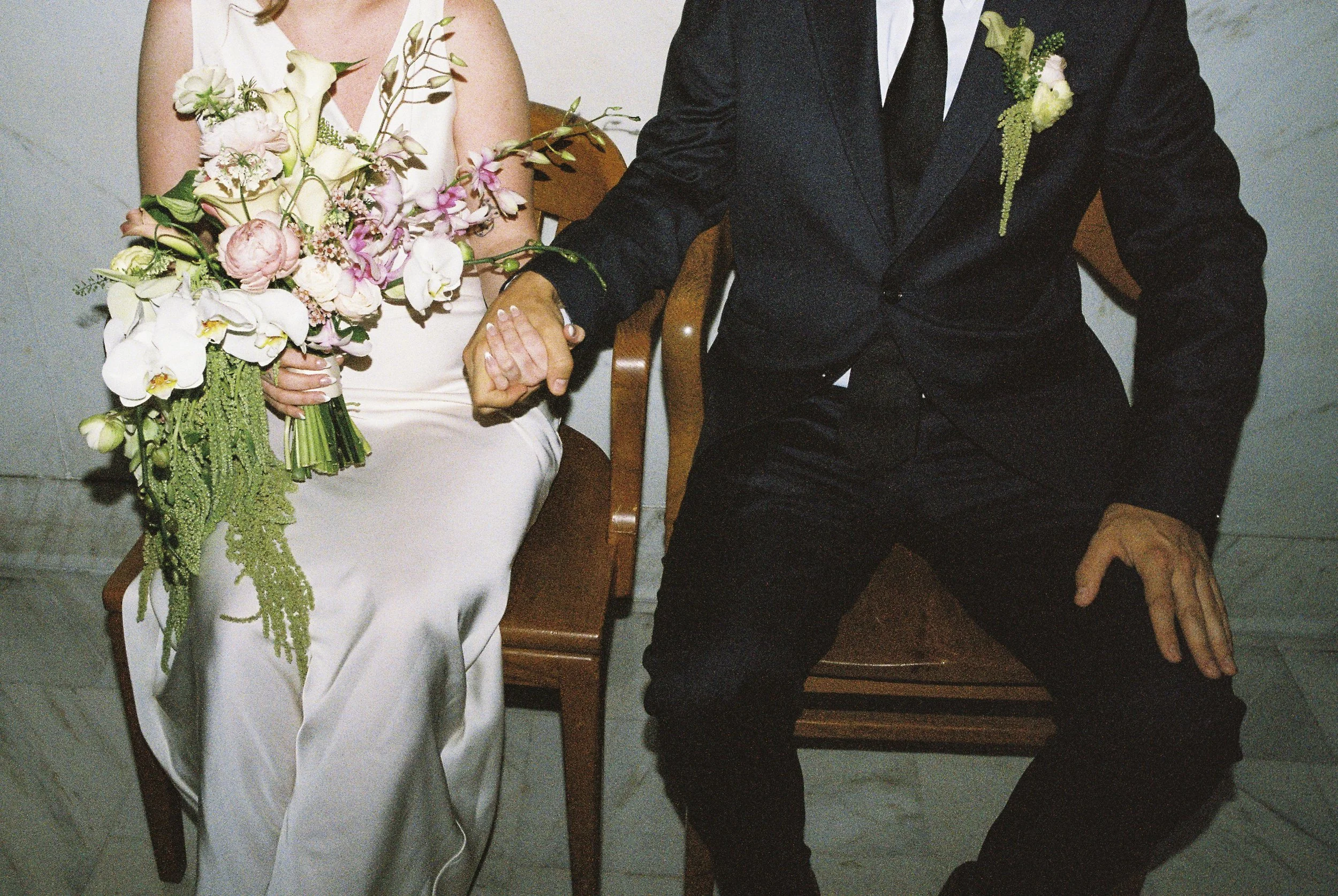 A bride and groom sitting closely, holding hands. The bride wears a white dress and holds a large bouquet of pink and white flowers with greenery. The groom wears a black suit with a white shirt and black tie, with a small floral boutonniere.