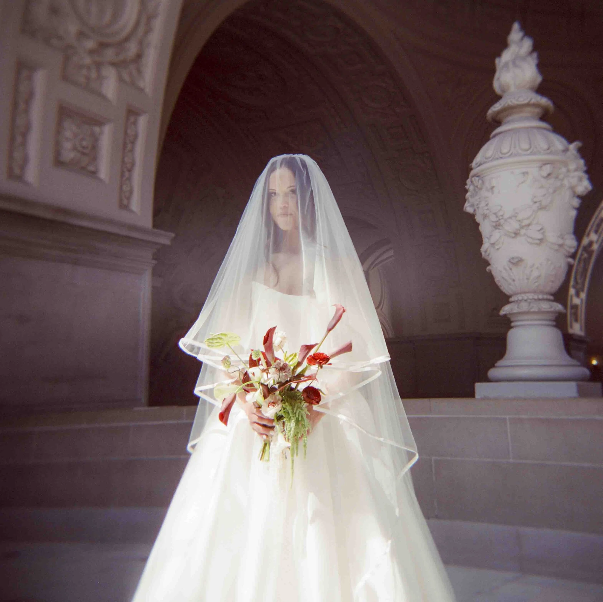 A woman dressed in a wedding gown holding a bouquet of flowers, standing indoors with ornate architectural details and a large decorative urn in the background, wearing a sheer white veil.