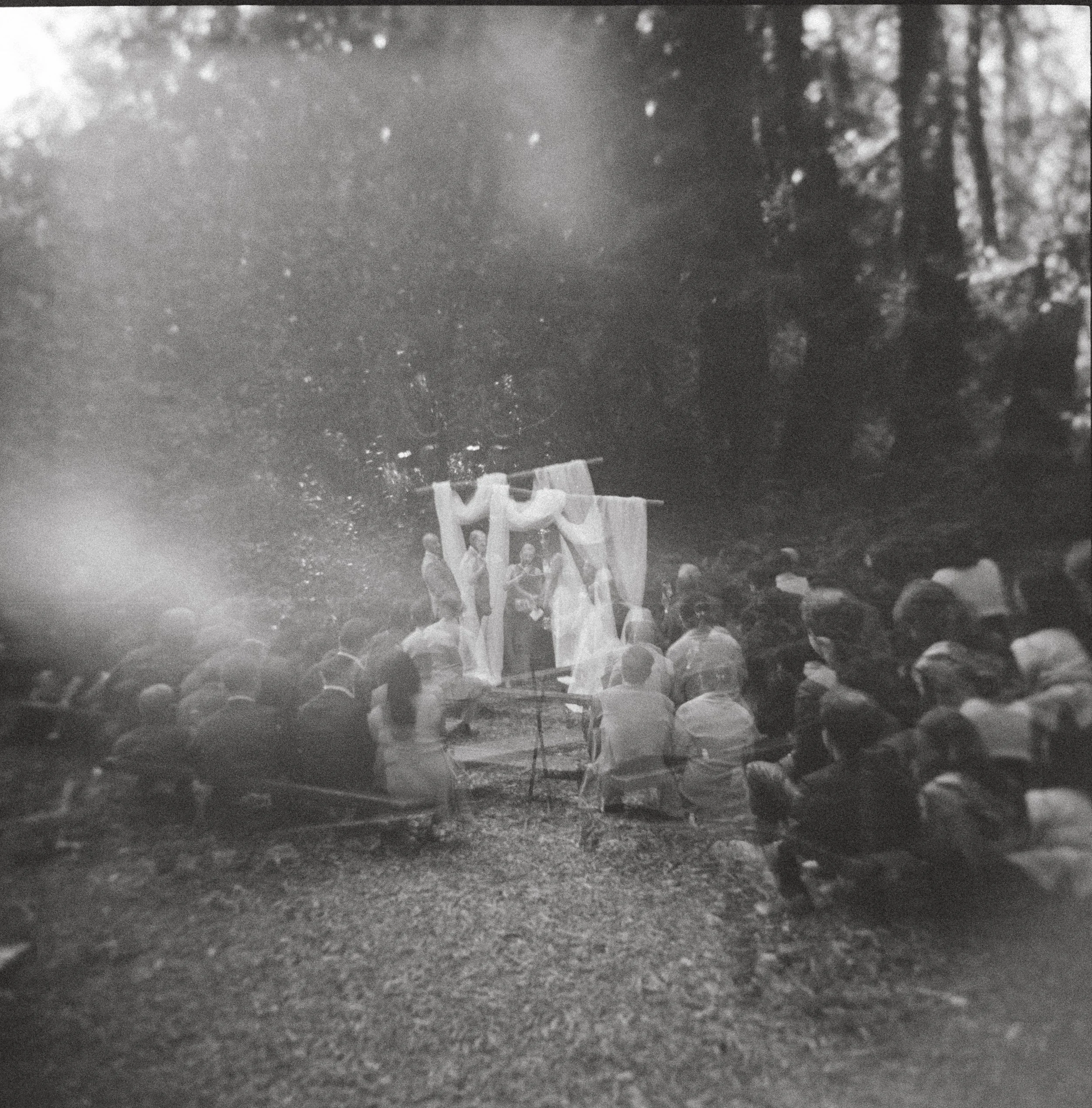 Black and white photo of an outdoor wedding ceremony with guests seated, facing the altar decorated with drapery, set in a wooded area.