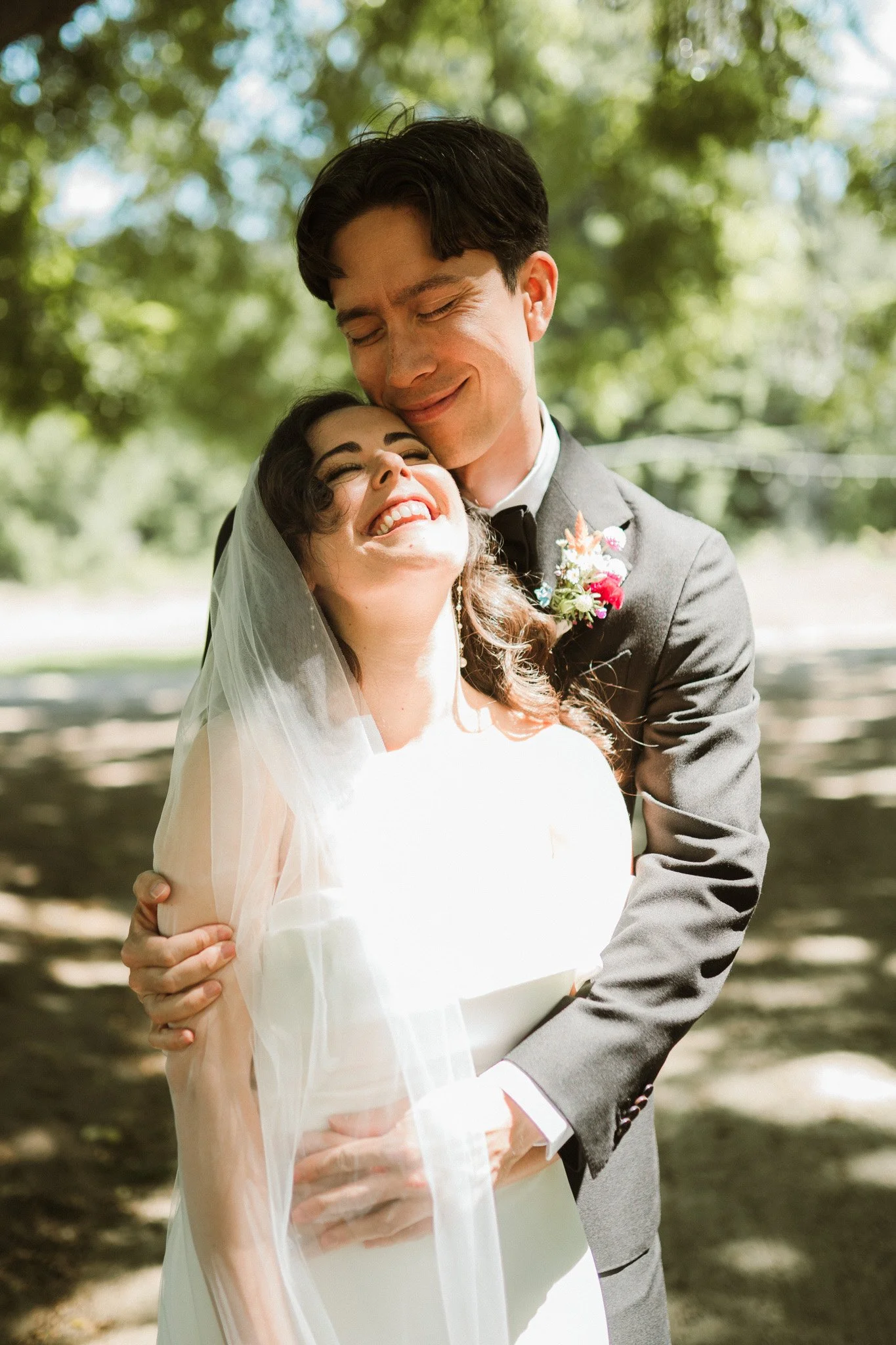 Bride and groom embrace, smiling, outdoors under a tree.