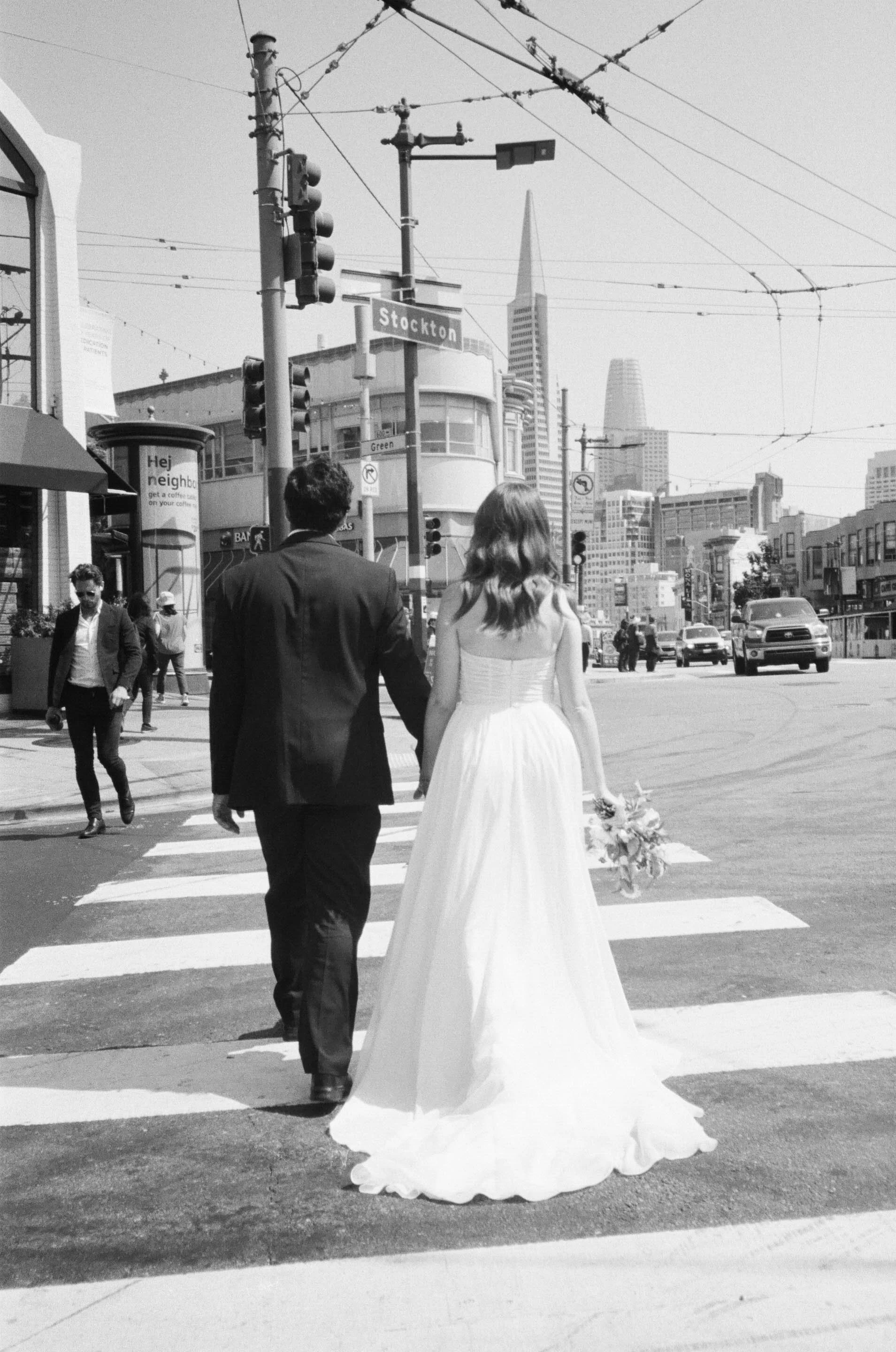 A couple in wedding attire crossing a street in a city, with the bride holding a bouquet. Urban buildings and traffic lights are visible in the background.