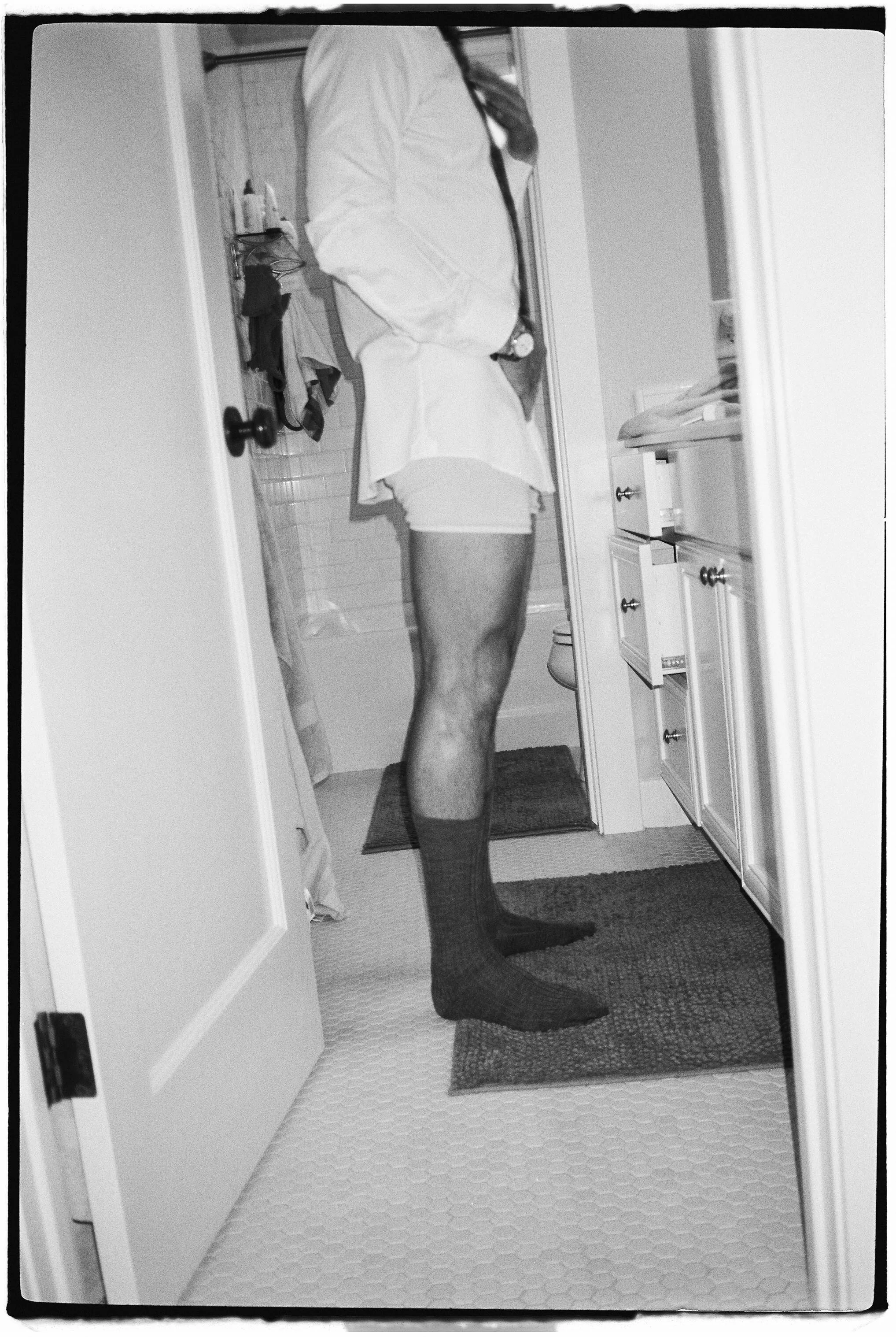 A black-and-white photo of a person taking a mirror selfie in a bathroom, wearing a blazer, shorts, and knee-high socks.