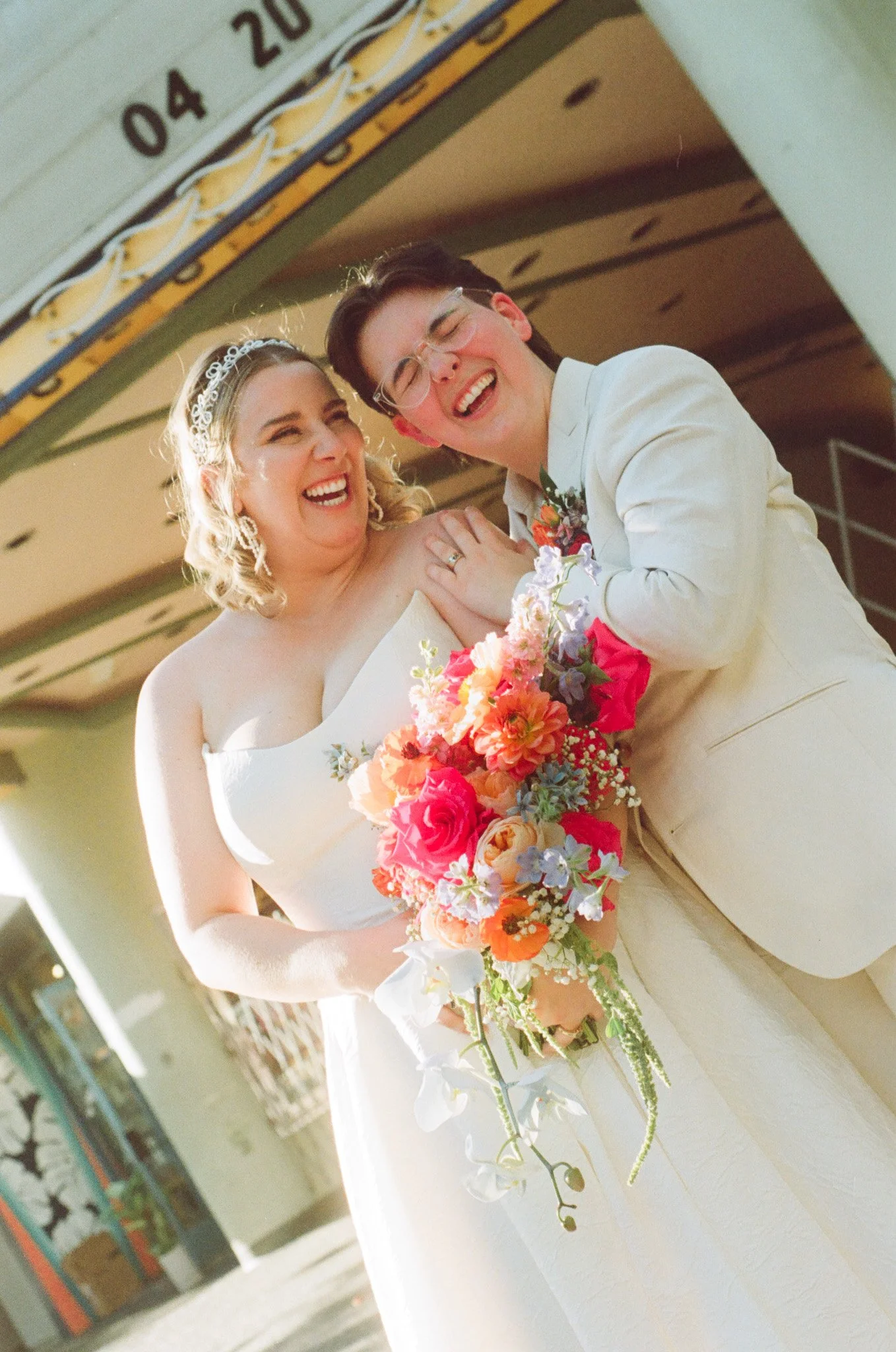 Two people dressed in wedding attire, smiling and holding a colorful bouquet with bright flowers.
