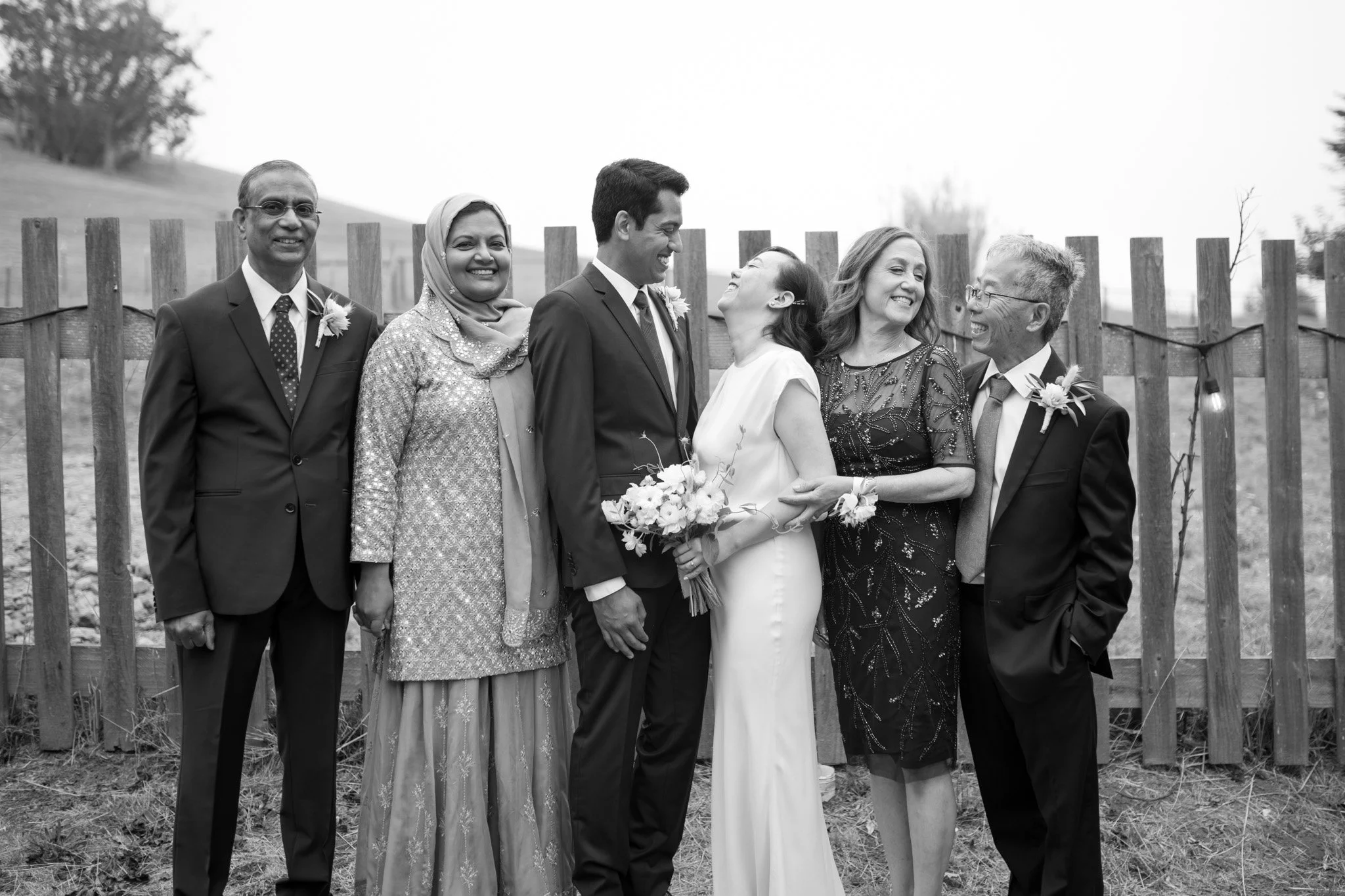 Group of seven people at a wedding, standing outdoors in front of a wooden fence, smiling and looking at each other, black and white photo.