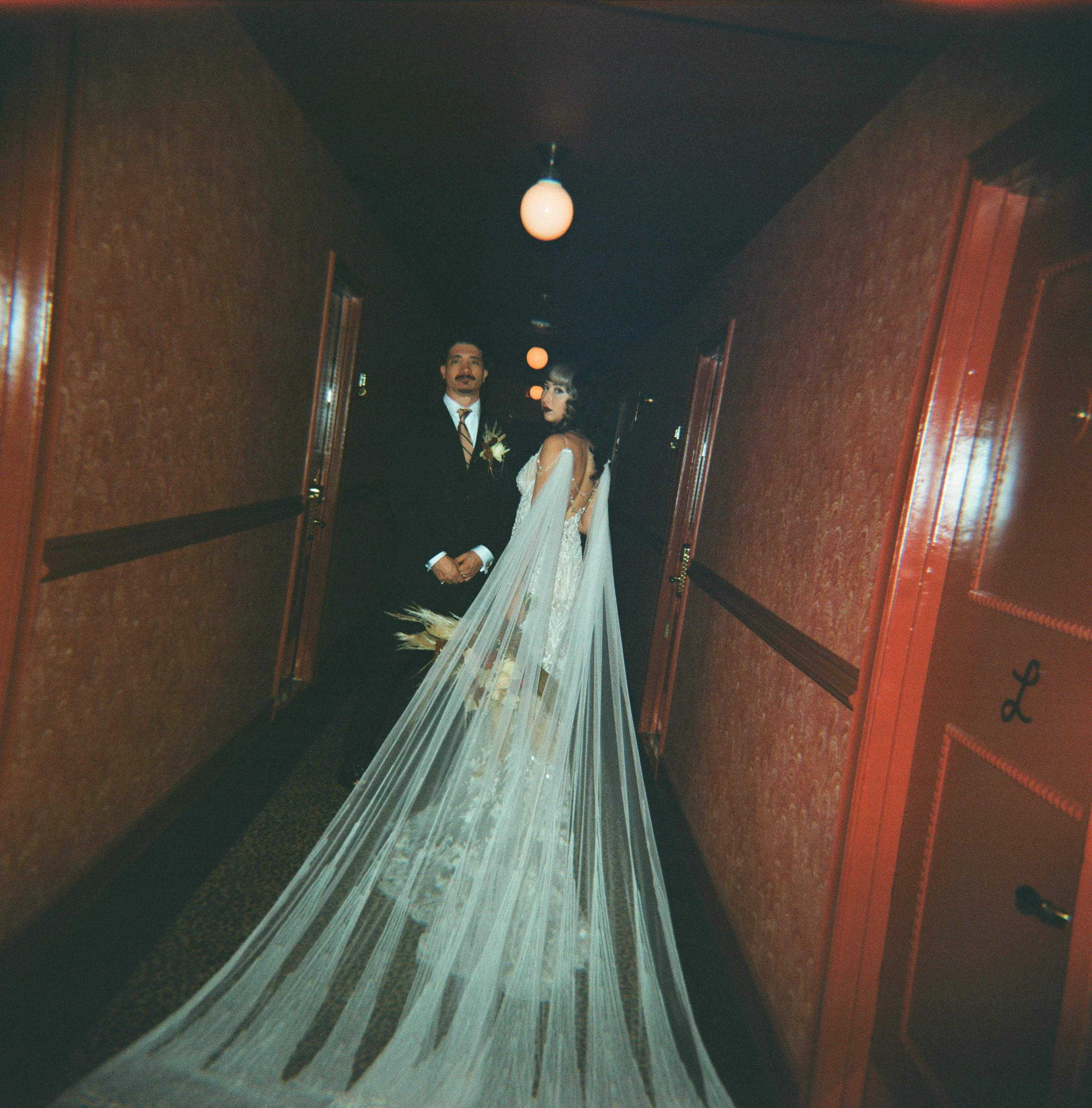 Bride and groom standing in a dimly lit hallway with the bride wearing a long veil.