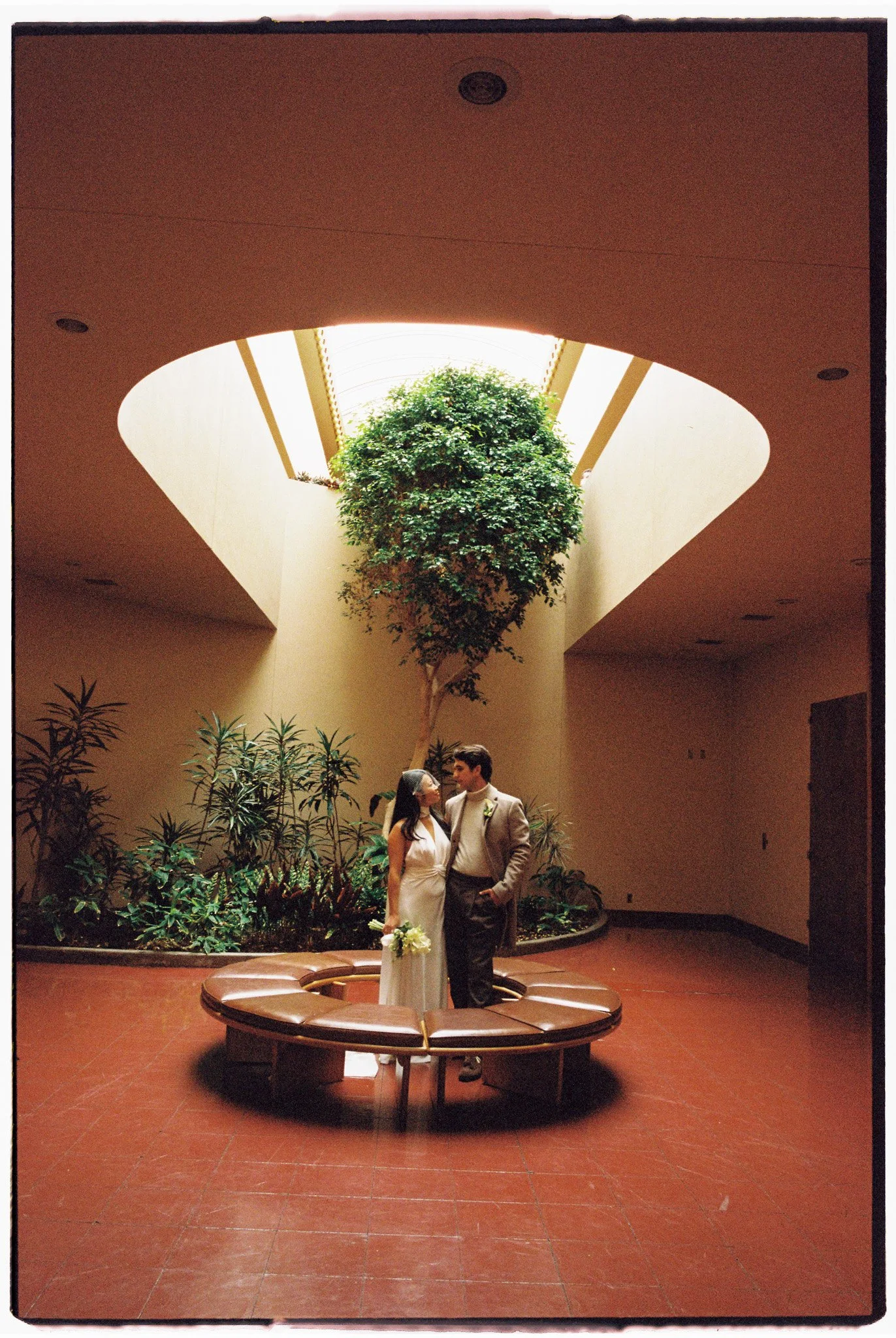 A couple stands together in the middle of an indoor courtyard. They are beside a circular bench, under a skylight with a large tree in the center. The setting is warmly lit, with plants lining the walls.