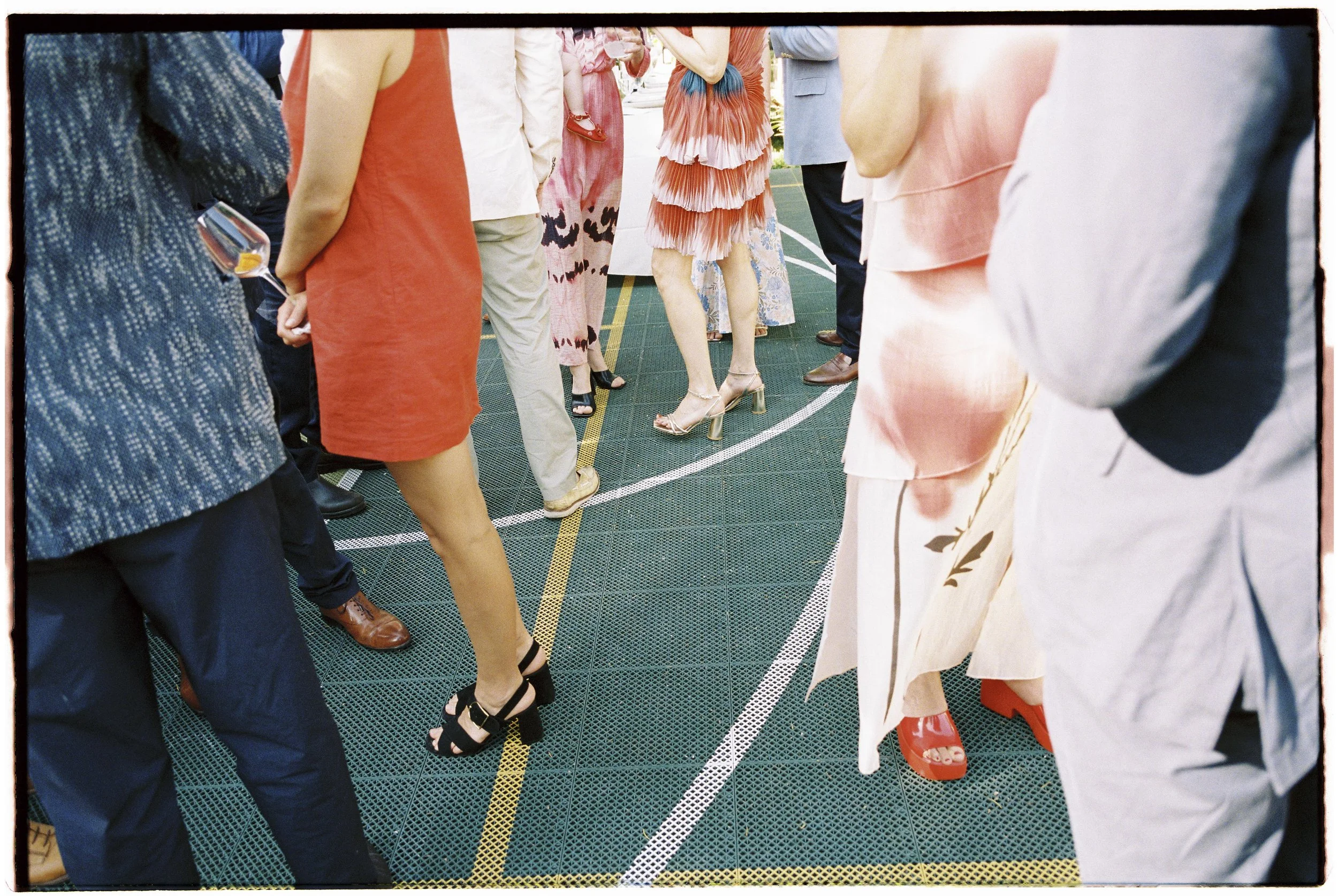 People wearing formal and colorful attire gathered on a green outdoor court, with only their legs and shoes visible.