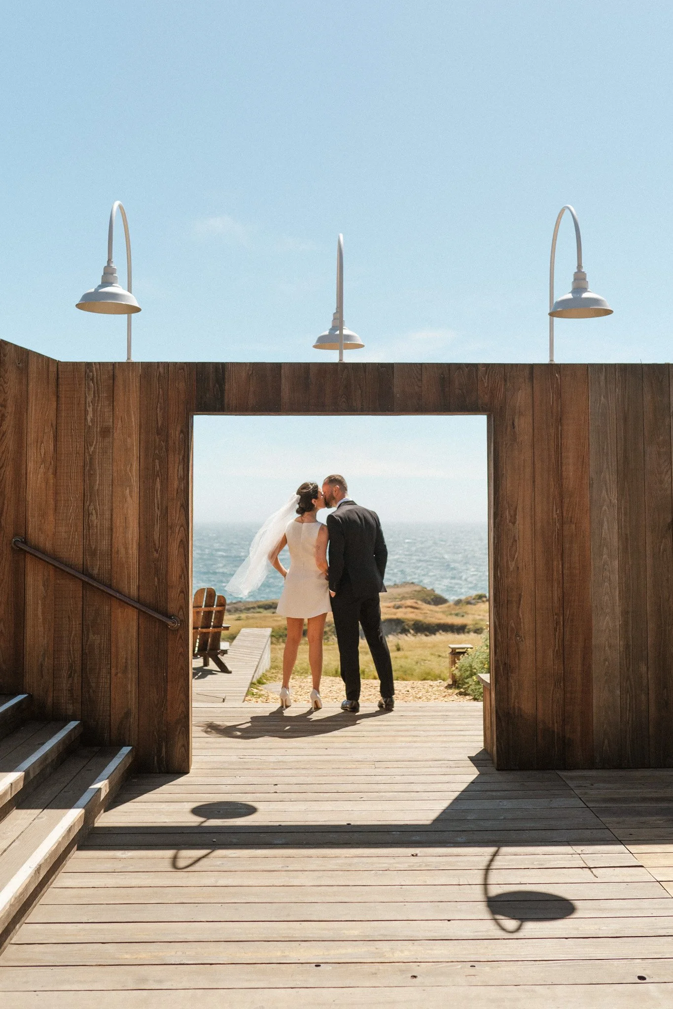 A bride and groom kissing outdoors near the ocean, framed by a wooden archway, with blue sky and water in the background.