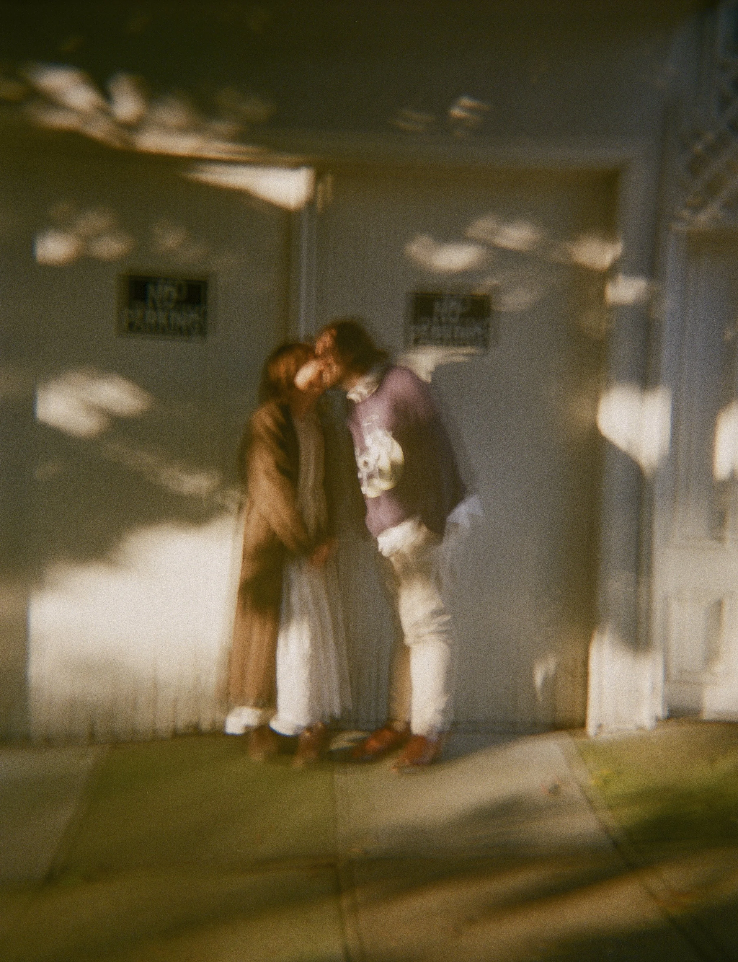 Two people standing close together, their foreheads touching, in front of a wall with 'No Parking' signs, with shadows and light reflections creating an abstract effect.