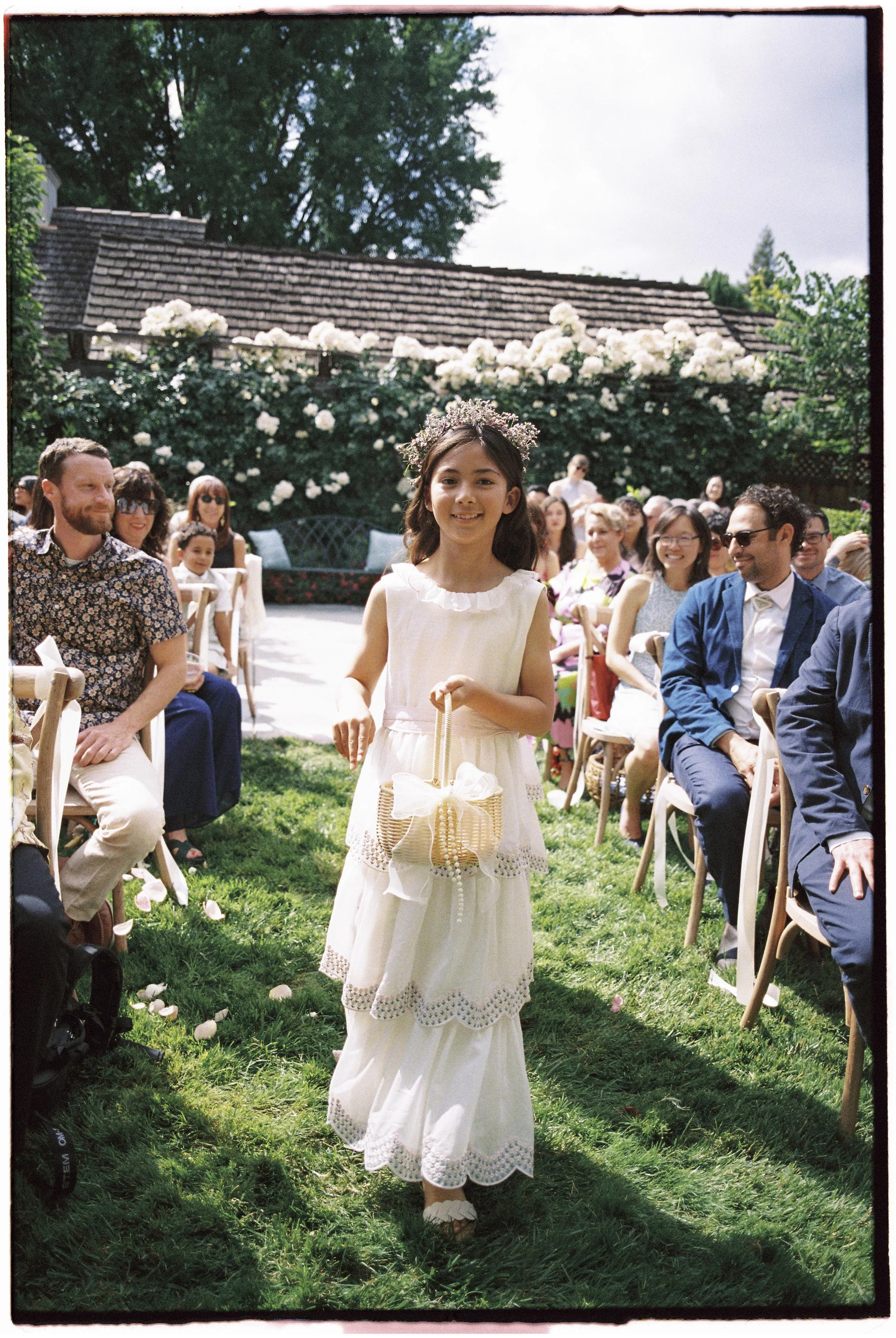 A young girl in a white dress walking down an outdoor aisle during a wedding ceremony, holding a basket and wearing a floral headpiece. Guests are seated on either side, smiling and watching her.