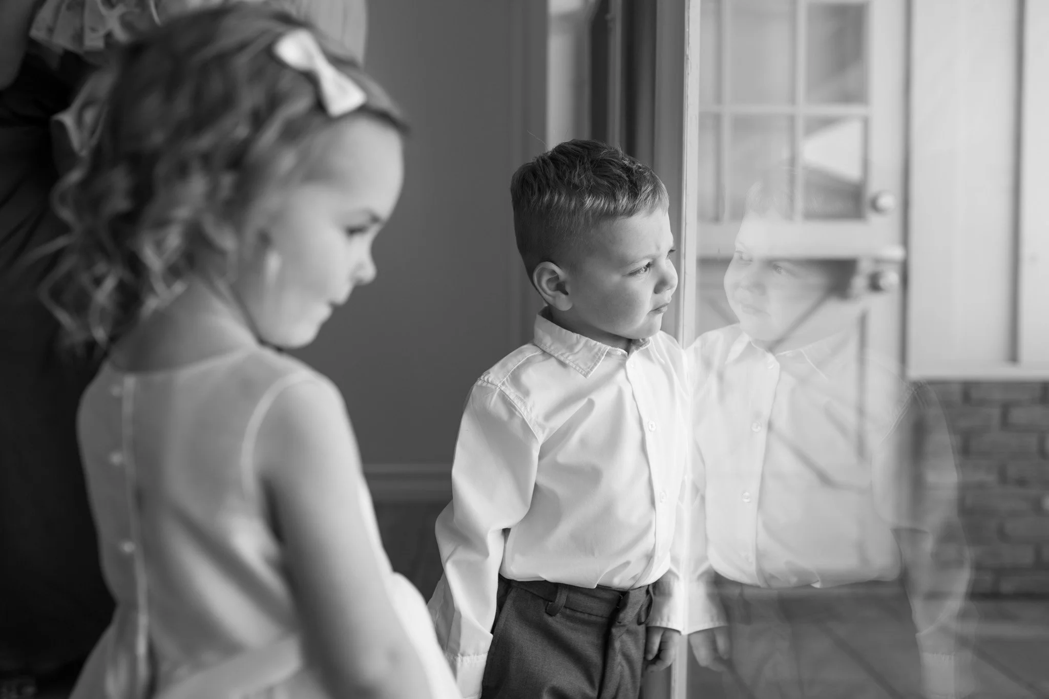A young girl and a boy standing near a window, looking outside, with the boy's reflection visible on the window.
