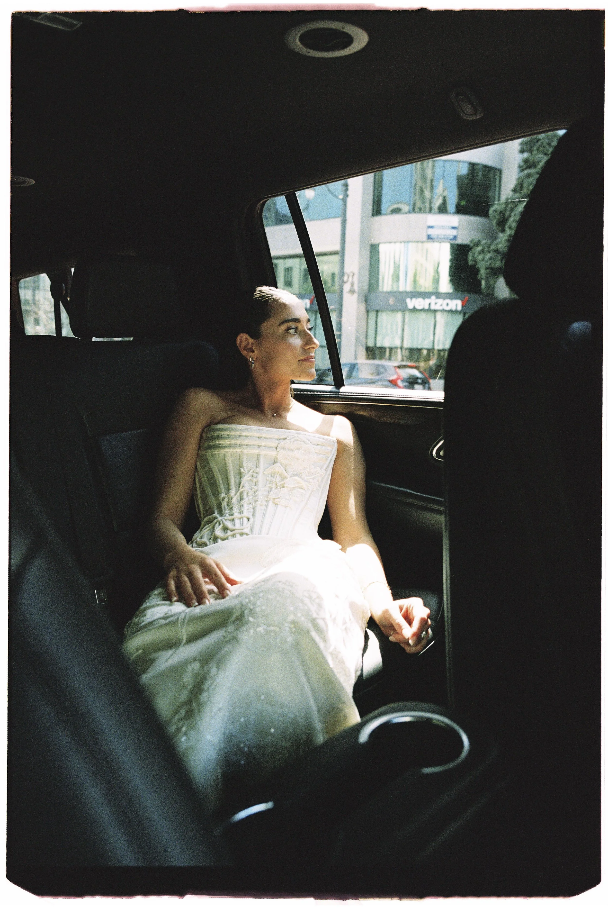A woman in a wedding dress sitting in the backseat of a car, looking out the window.