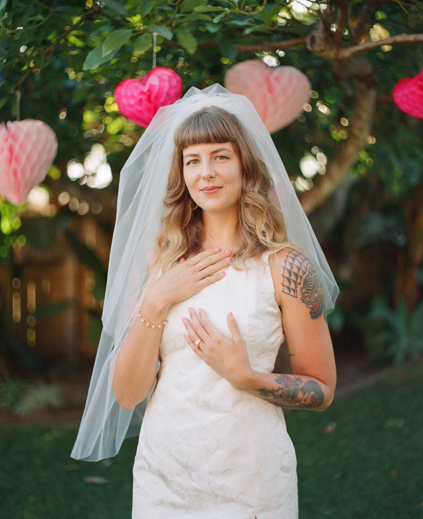 Bride in a wedding dress with tattoos, standing outdoors, with pink heart decorations in the background.