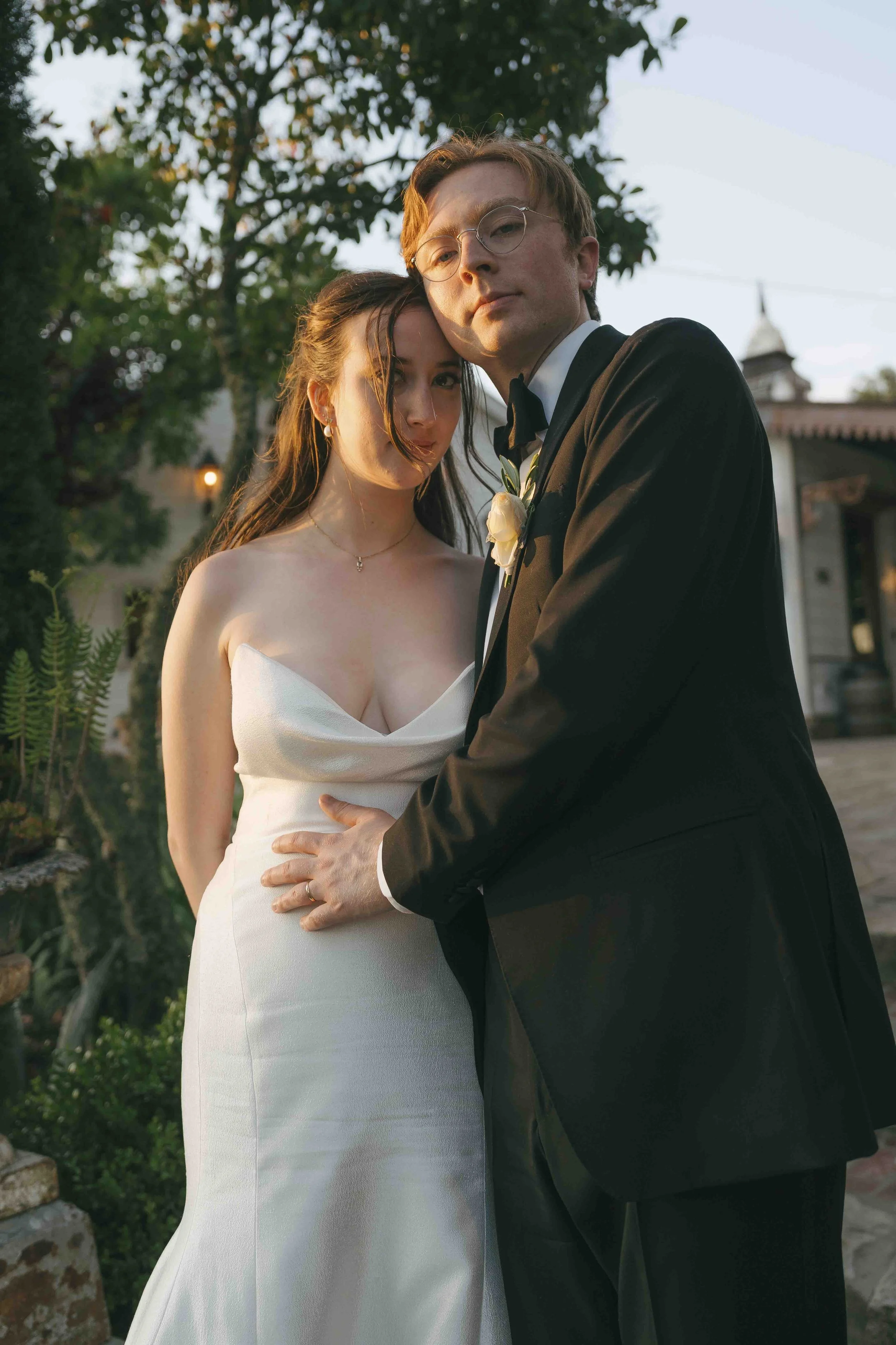 A bride and groom pose in a garden during sunset. The bride wears a strapless white wedding dress and a delicate necklace, while the groom is in a black tuxedo with a boutonniere. They stand close with the groom's hand on the bride's waist, both look