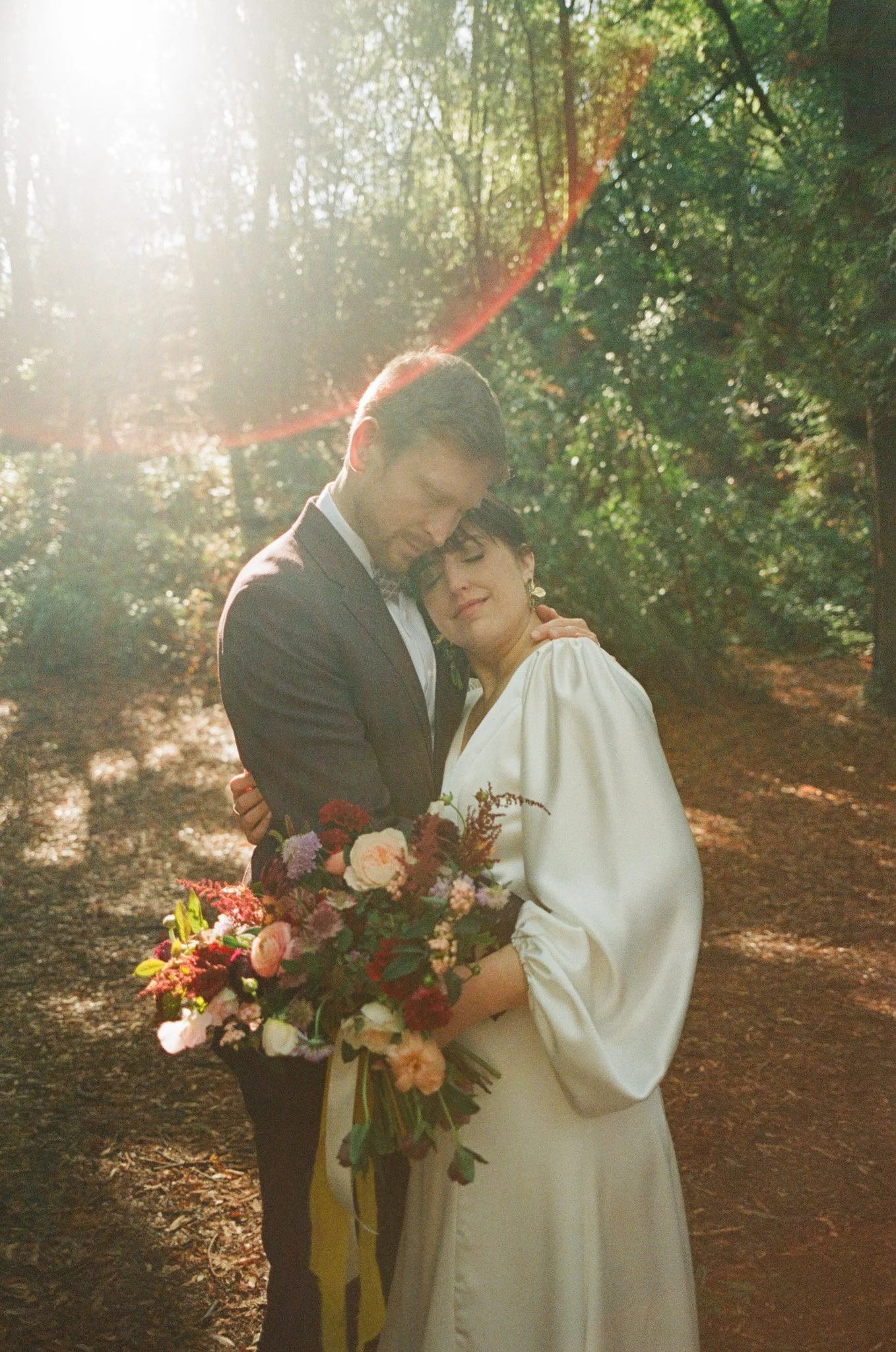 A couple dressed in wedding attire hugging in a forest with sunlight shining through trees, holding a bouquet of flowers.