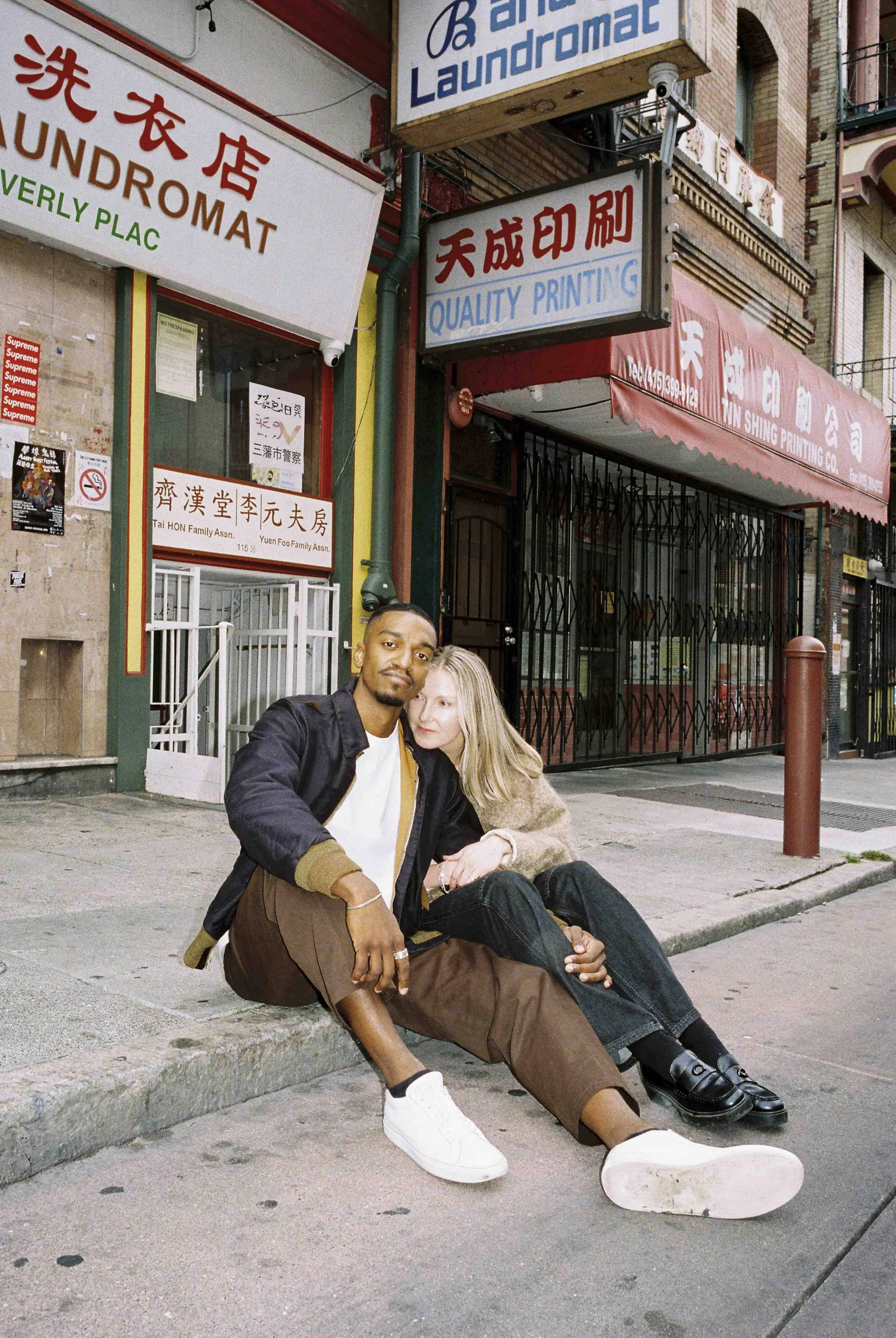A young man and woman sitting on a city sidewalk at night, with signs in Chinese and English for laundromat, printing, and other businesses visible behind them.