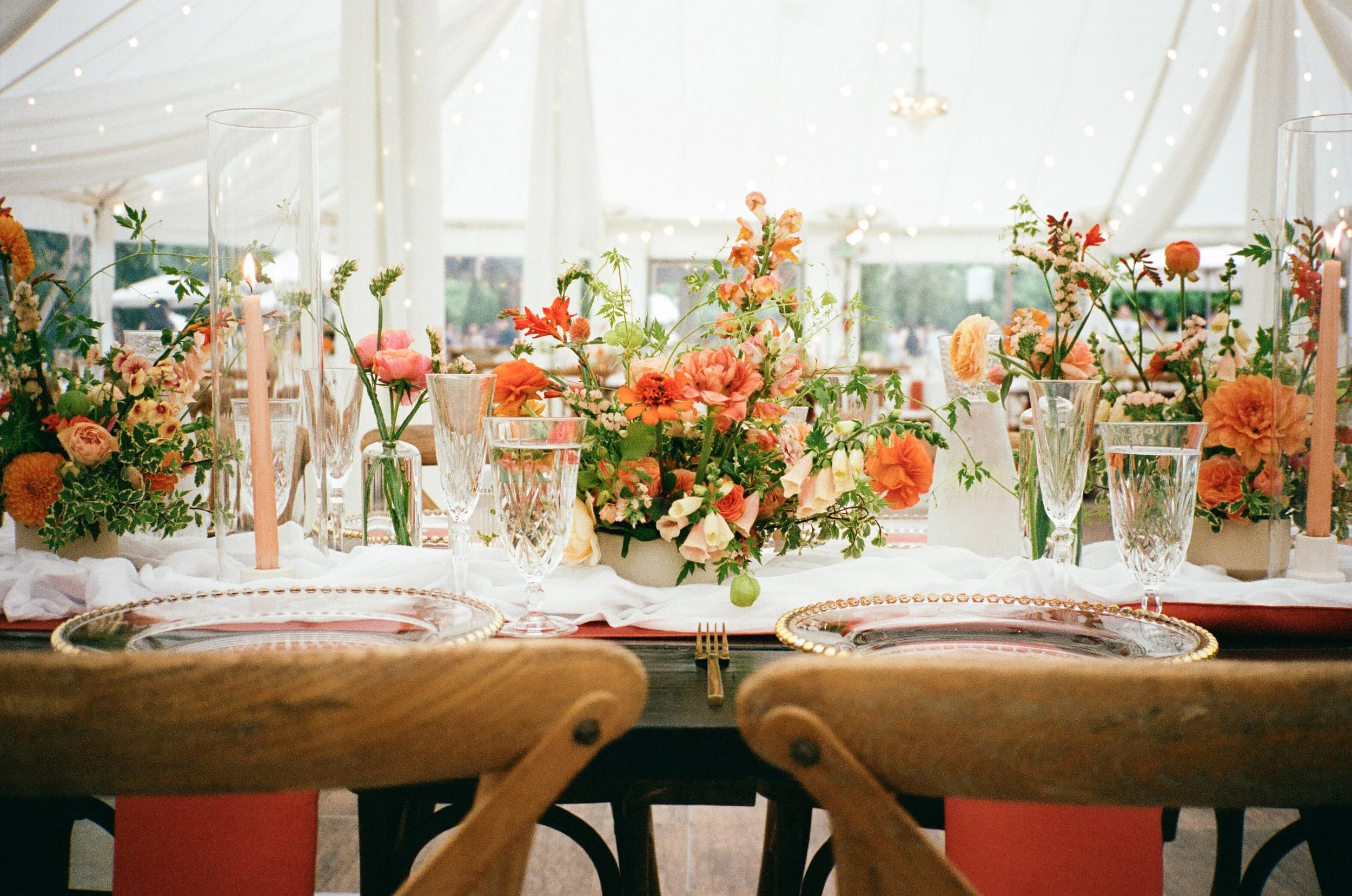 Elegant table setting with floral centerpieces, gold-rimmed plates, crystal glassware, and tall candles under a white tent.