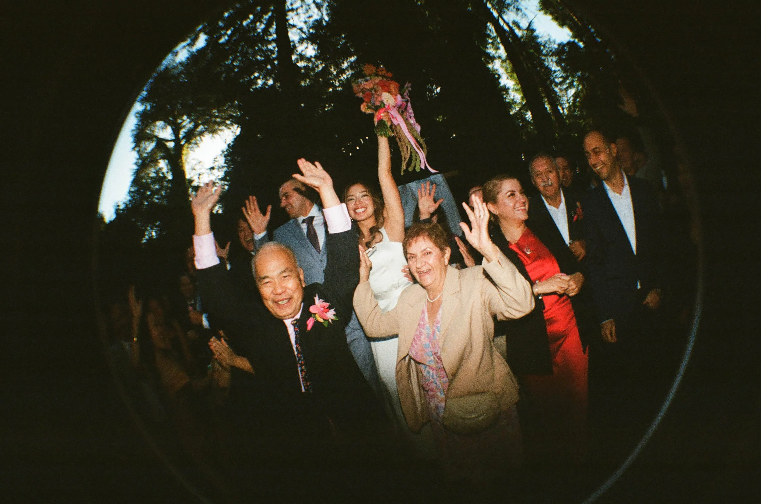 People celebrating at a wedding, some with raised hands, a woman holding a bouquet of flowers, and joyful expressions, outdoors among tall trees.