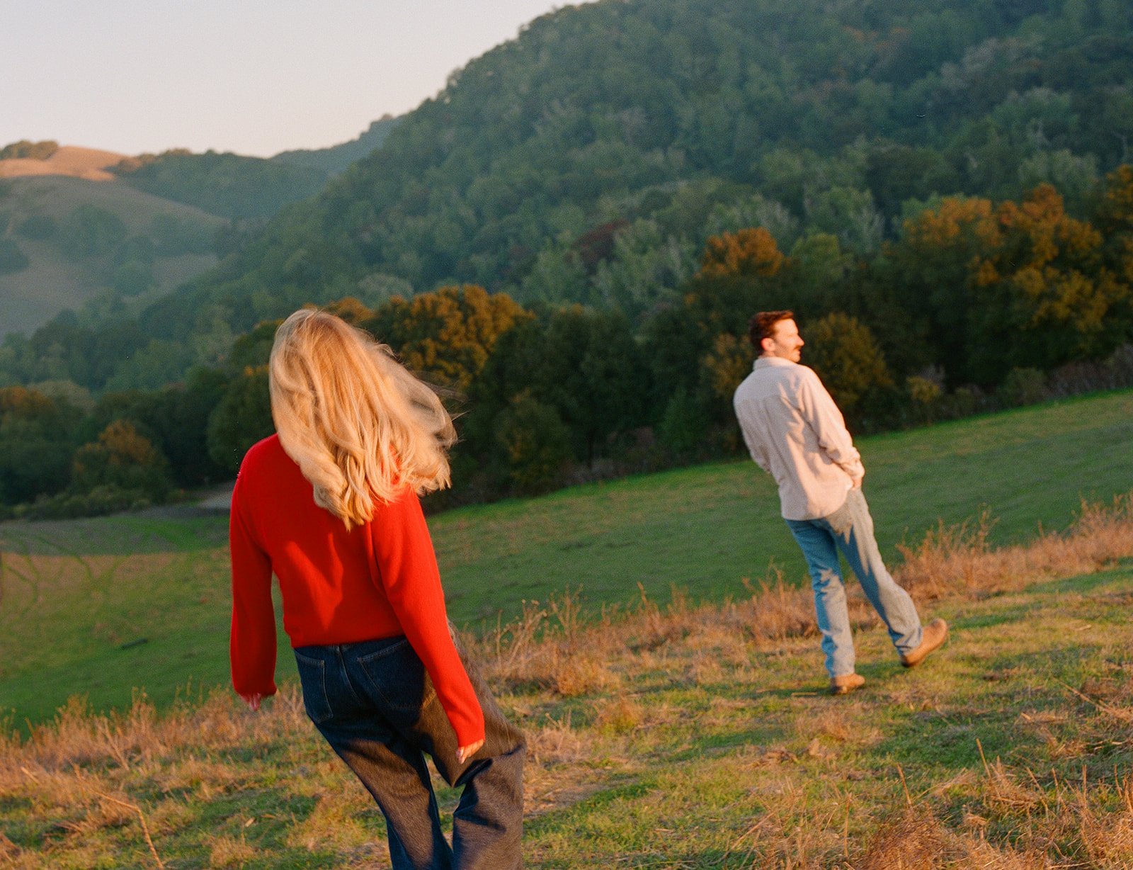 A woman with blonde hair in a red sweater and dark pants walking in a grassy field, with a man in a light-colored jacket and jeans smiling and looking away in front of her, set against a backdrop of green hills and trees during sunset.