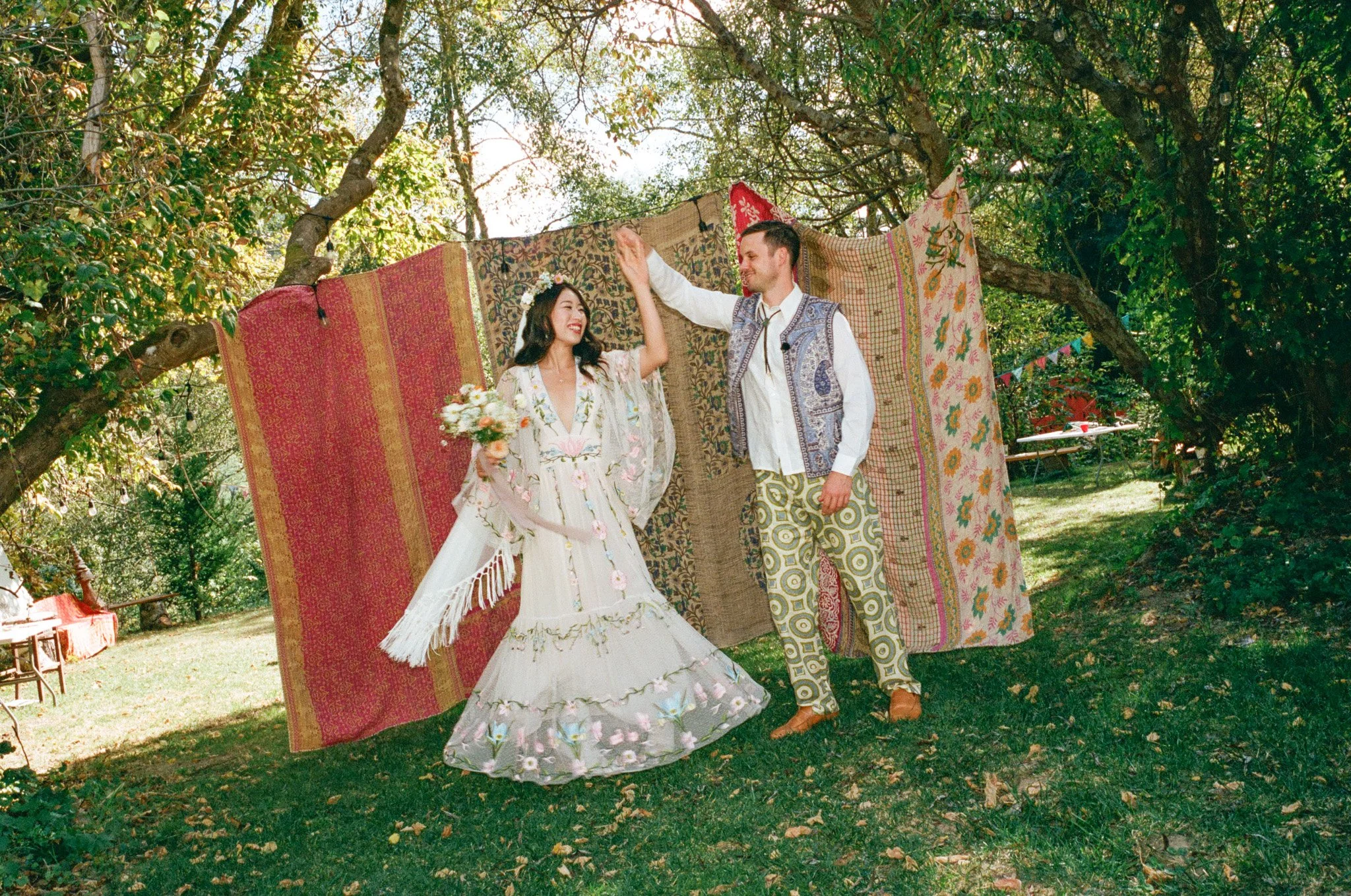 Bride and groom in a whimsical outdoor wedding with colorful tapestries and greenery.