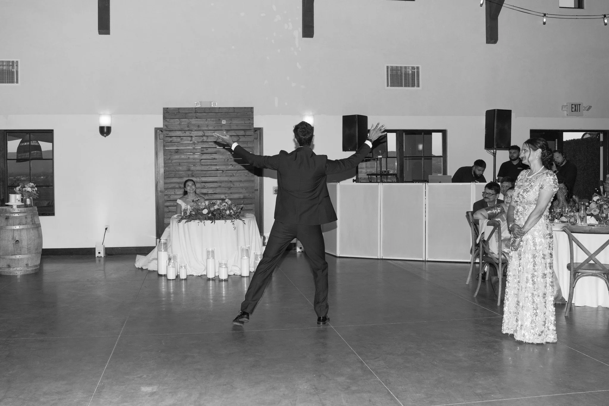 A man in a suit is dancing with arms outstretched in a reception hall. A woman in a lace dress is watching him. There are guests sitting at tables, a decorated table with flowers, and a DJ setup in the background.