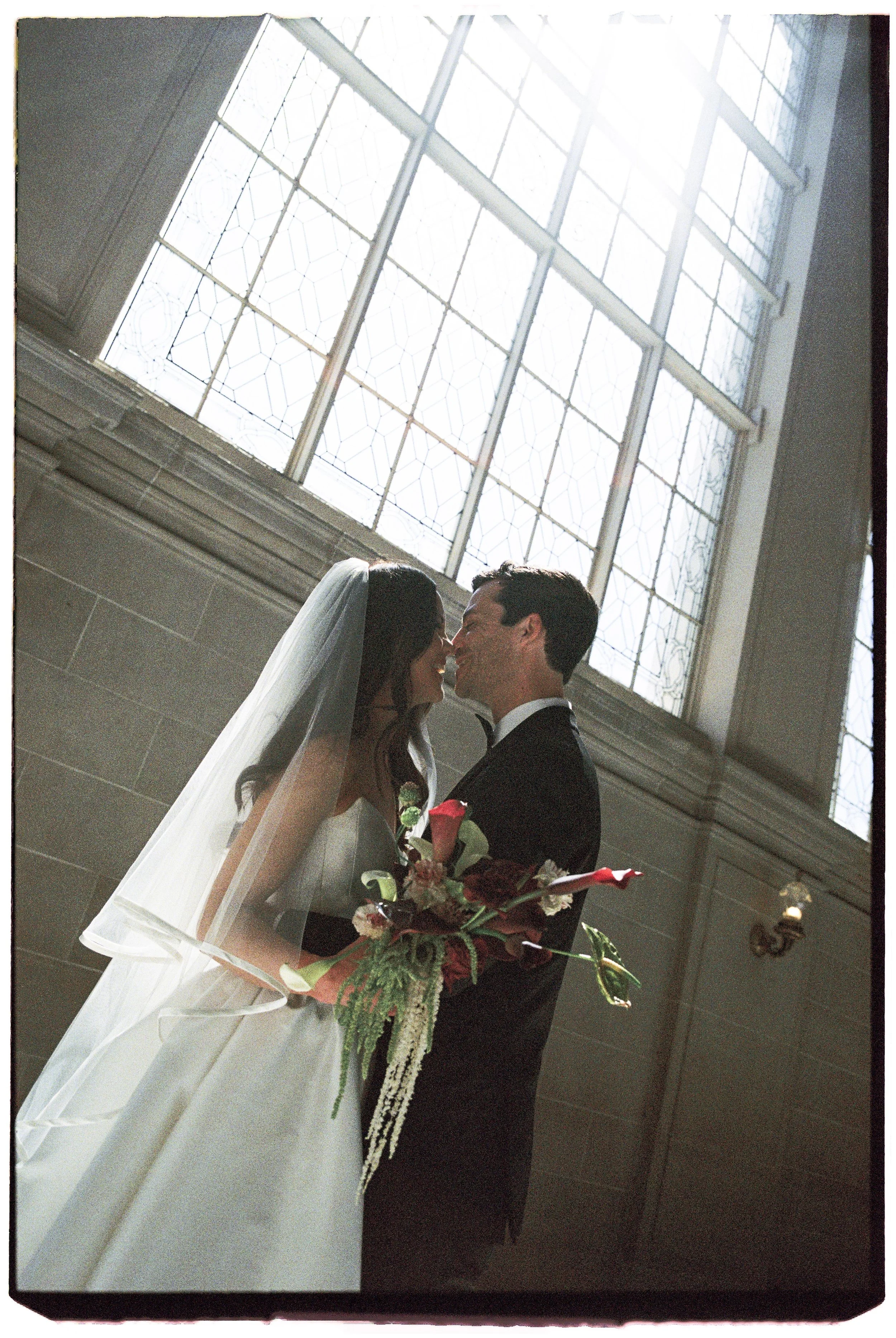 A bride and groom standing close together inside a church, holding a bouquet, with a large stained glass window behind them.