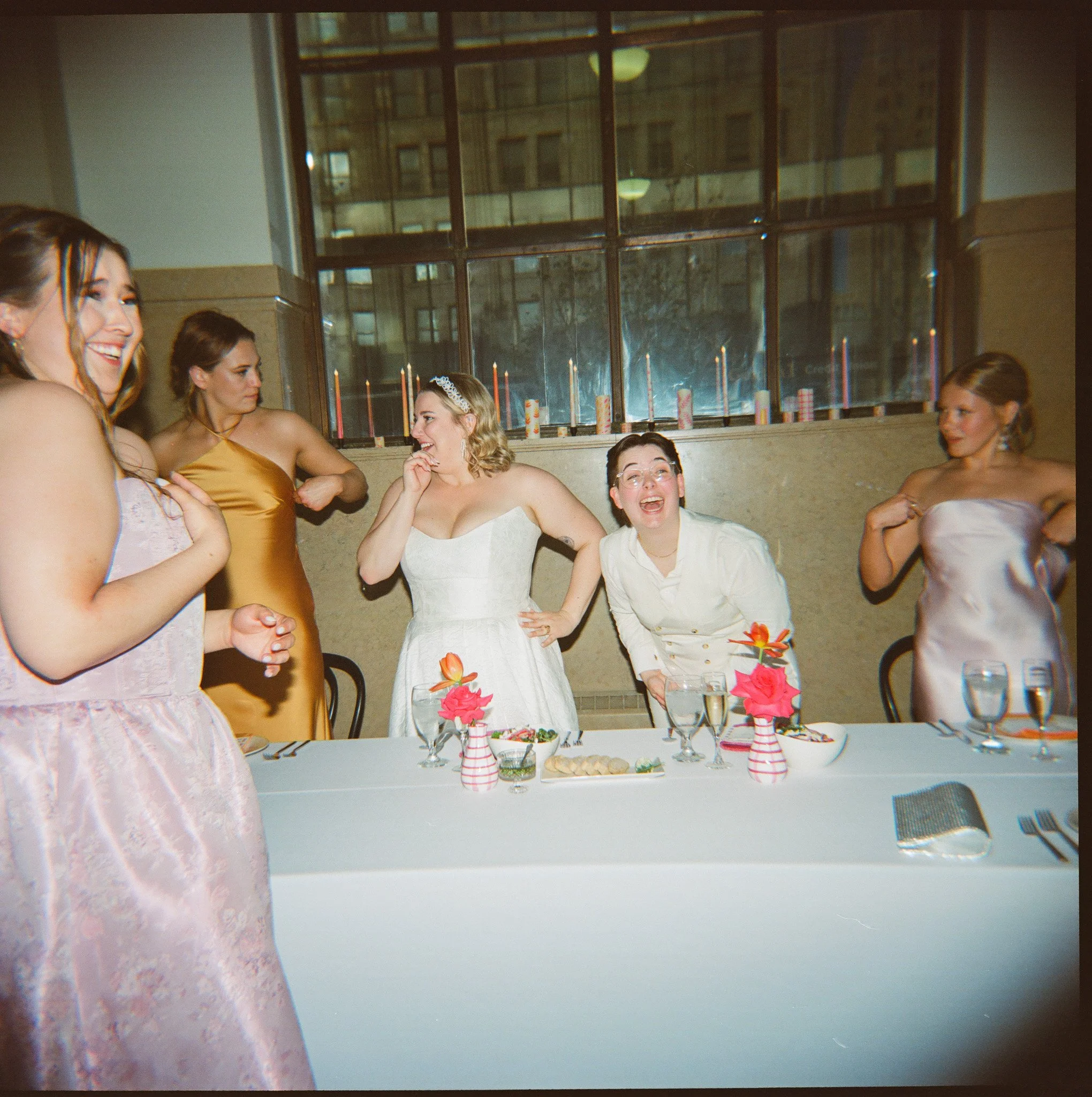 A group of people in formal attire laughing and posing near a table with flowers and dishes.