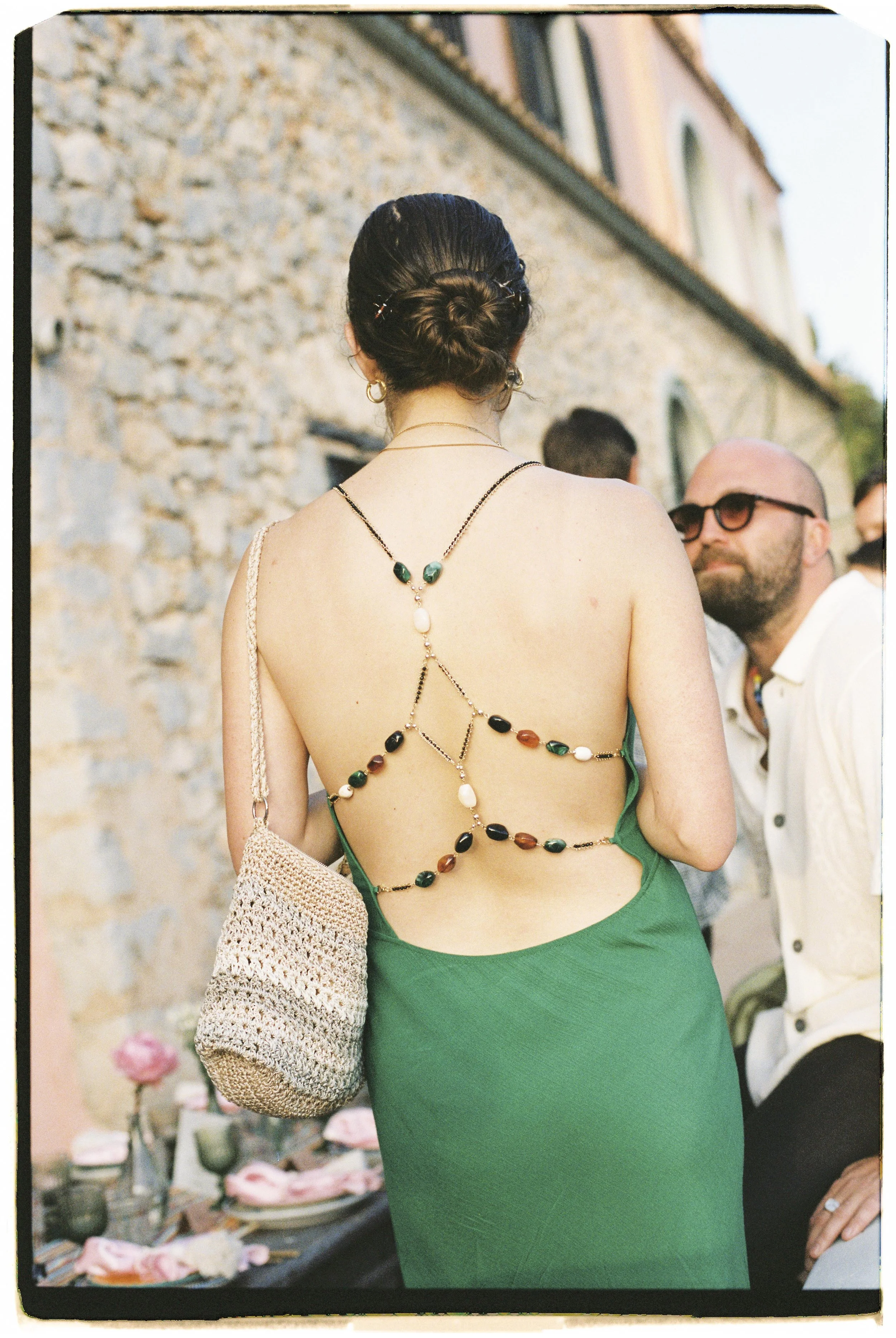 Back view of a woman wearing a green dress with an intricate beaded back chain, carrying a woven bag, at an outdoor event with a stone wall in the background and a man in glasses nearby.