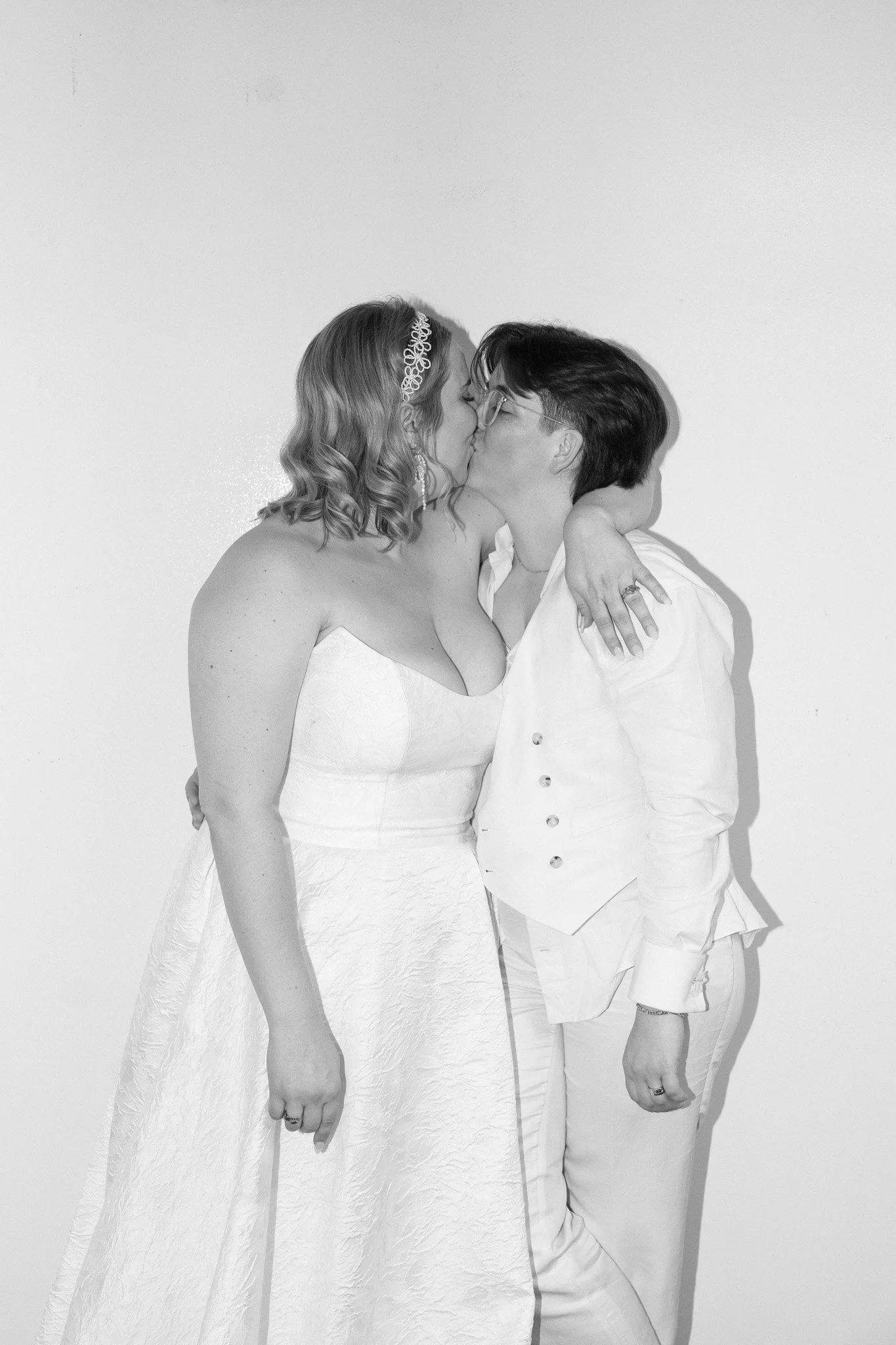 Black and white photo of two women kissing, one in a wedding dress and the other in white suit.
