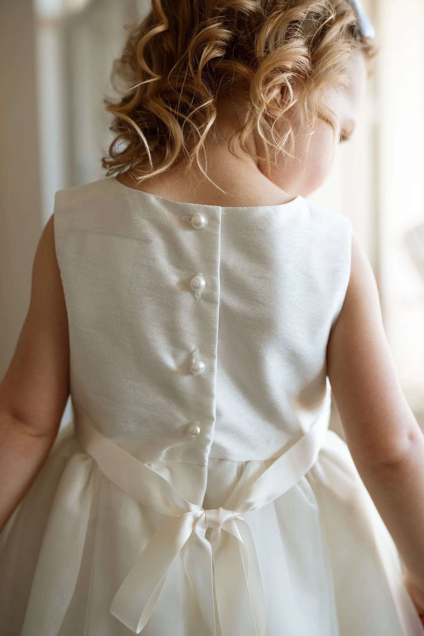A young girl in a white dress with pearl buttons and a satin sash tied in a bow, with blonde curly hair.