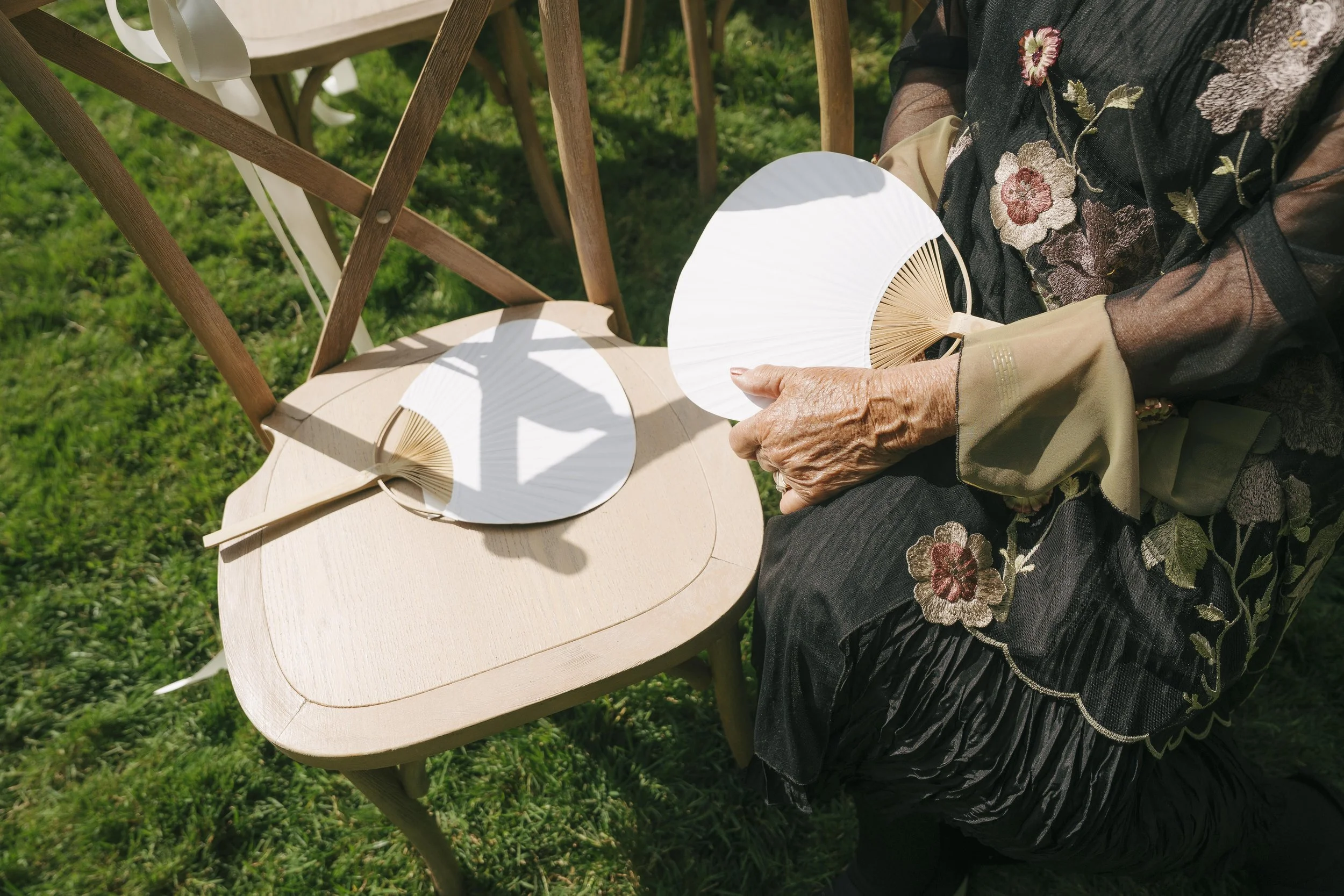 A person wearing a black floral dress sitting next to a light-colored wooden chair outdoors on grass, holding a white paper fan.