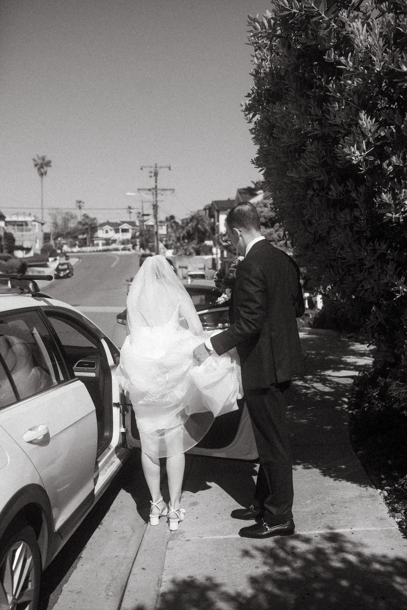 Bride and groom standing by a car on a street, bride wearing a veil and holding her dress, groom in a suit.