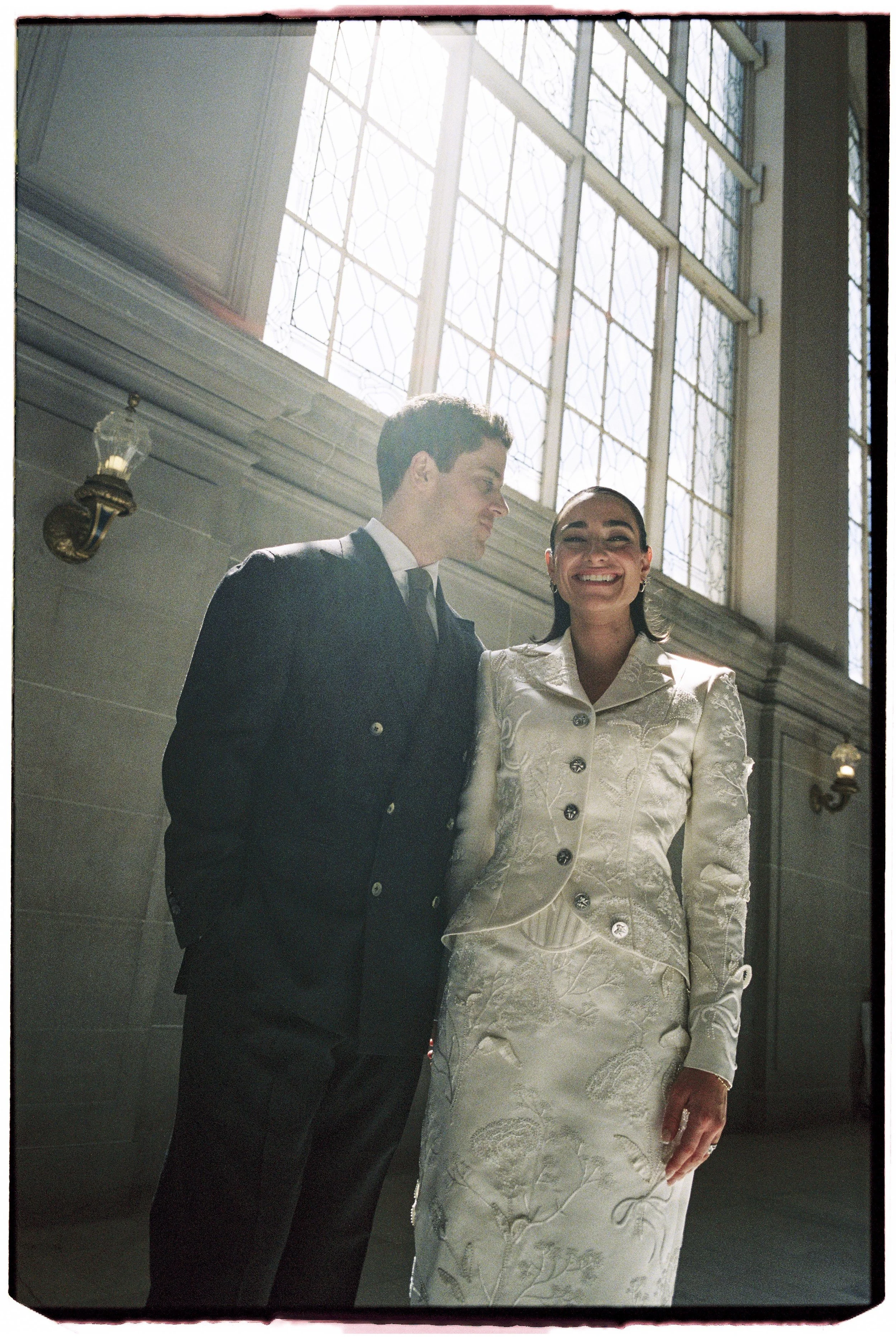 A man and a woman standing close together inside a building with large stained glass windows, the woman smiling brightly.