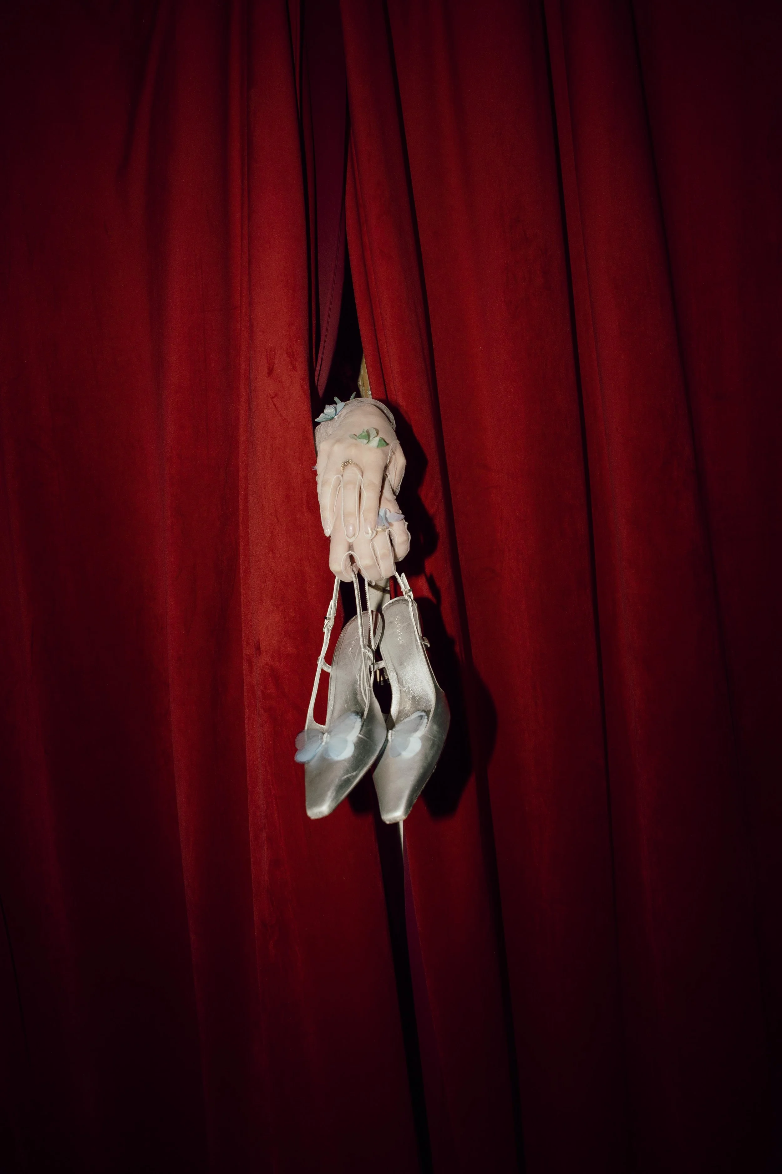 A hand holding a pair of silver high-heeled shoes with butterfly details, hanging against a red curtain background.