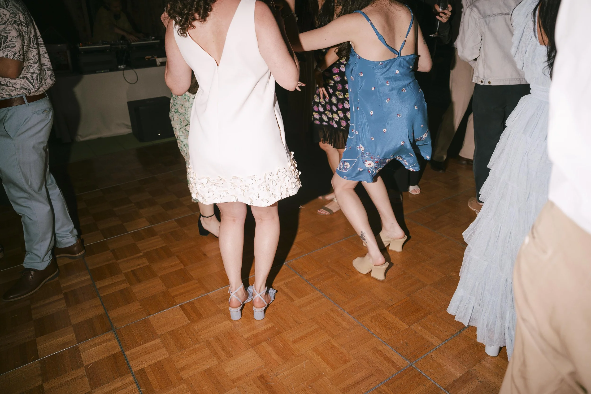People dancing on a wooden floor at a party, wearing various dresses and footwear.