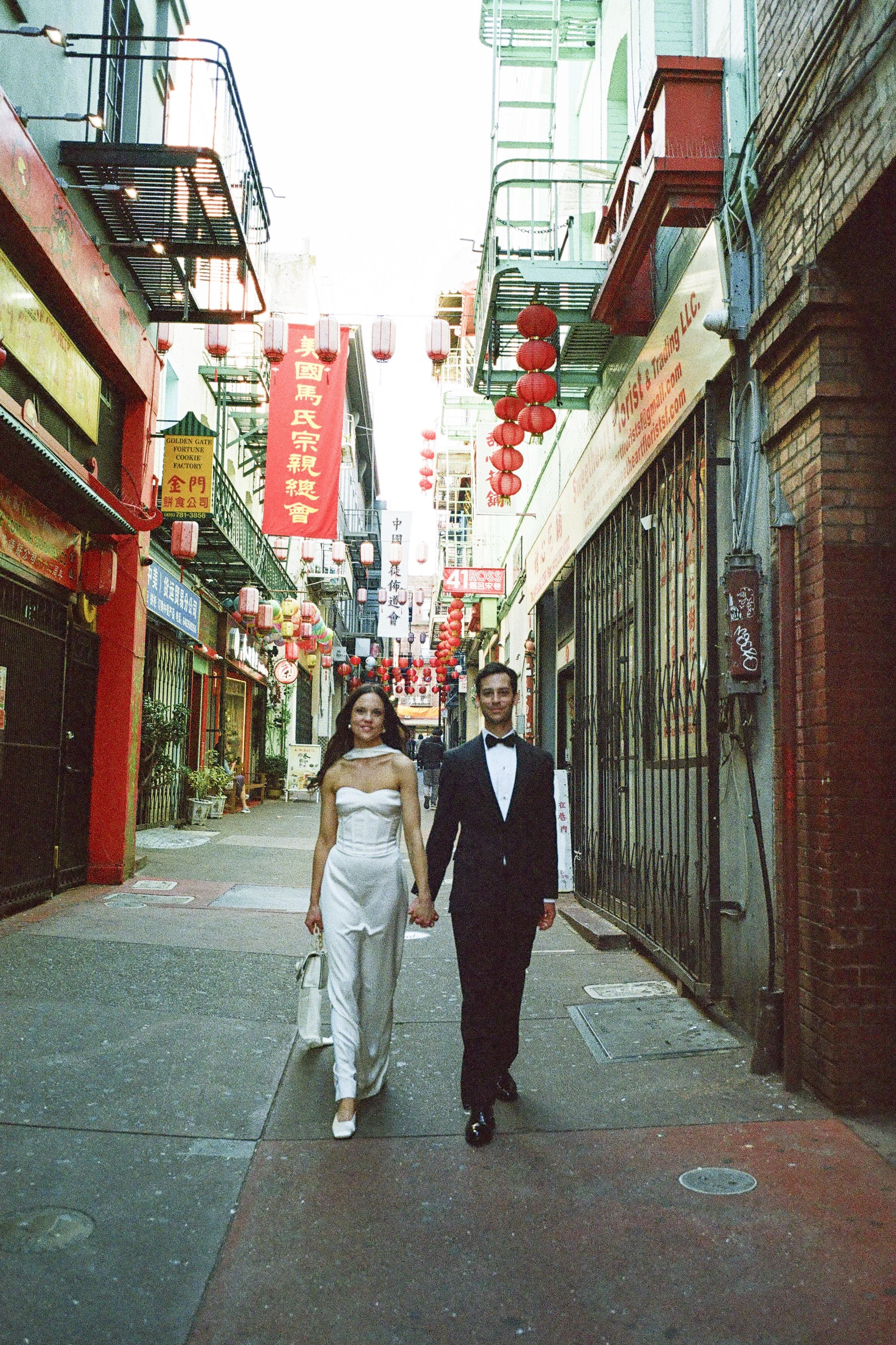 A couple dressed in formal wedding attire walking hand in hand down a narrow street decorated with red Chinese lanterns and banners.