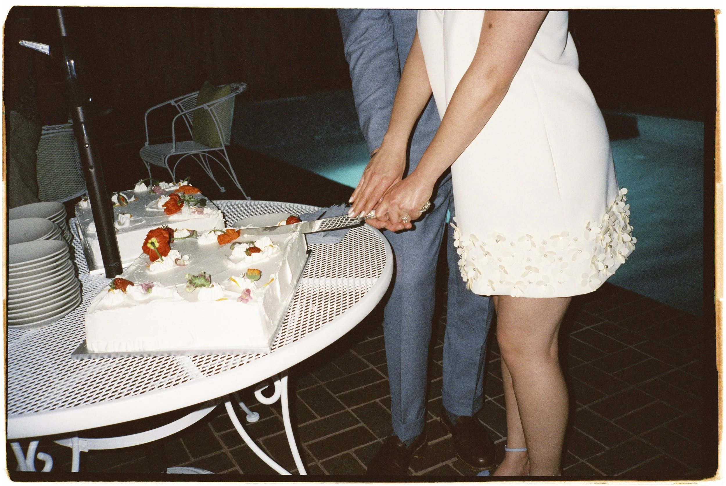 A couple is cutting a wedding cake together, with the bride wearing a white dress decorated with floral appliquΓ©s, and the groom wearing a gray suit. The wedding cake is rectangular, decorated with flowers, and placed on a white metal table.