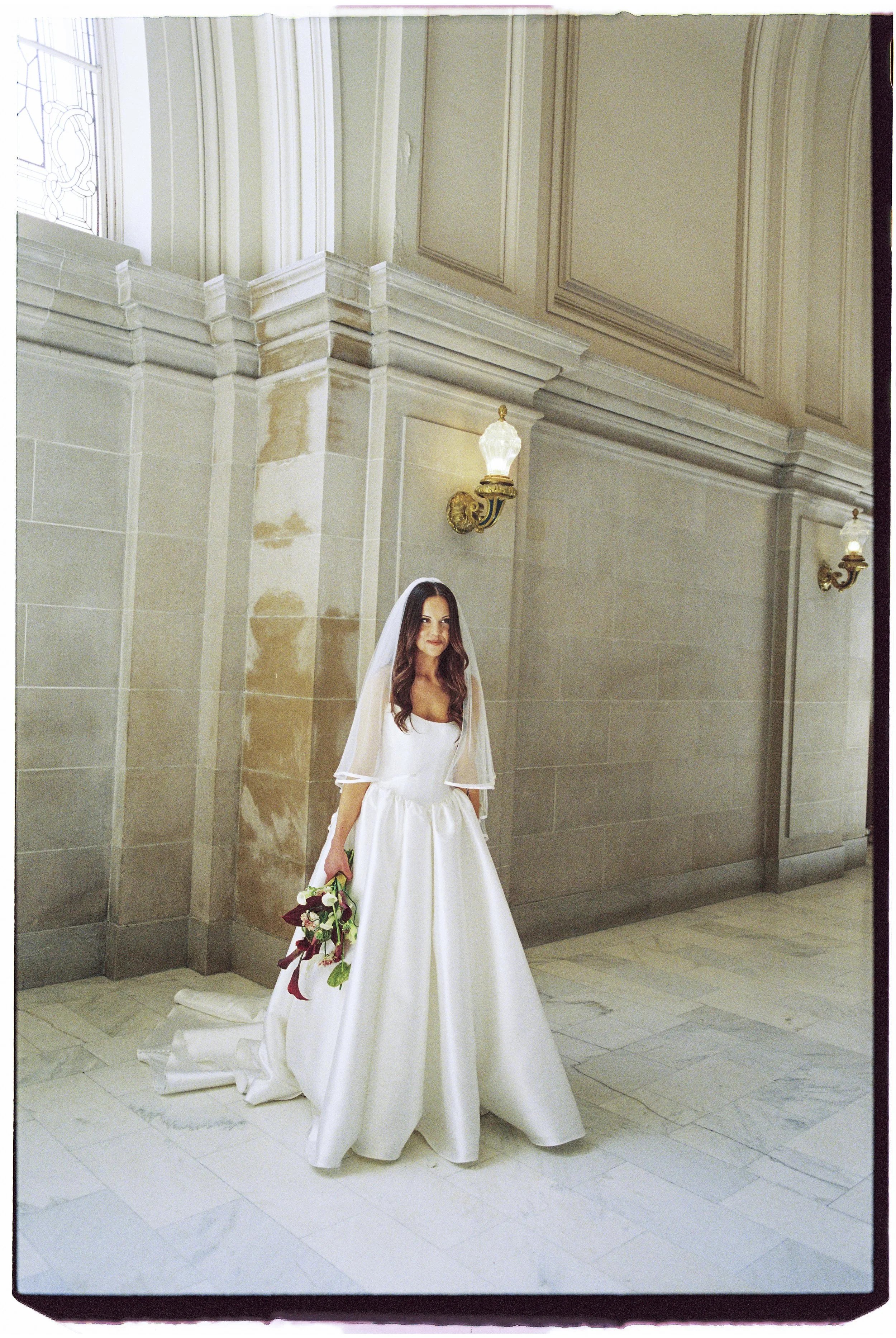 A bride in a white wedding dress and veil holding a bouquet, standing inside a grand, elegant building with marble floors and beige walls, illuminated by wall-mounted lamps.