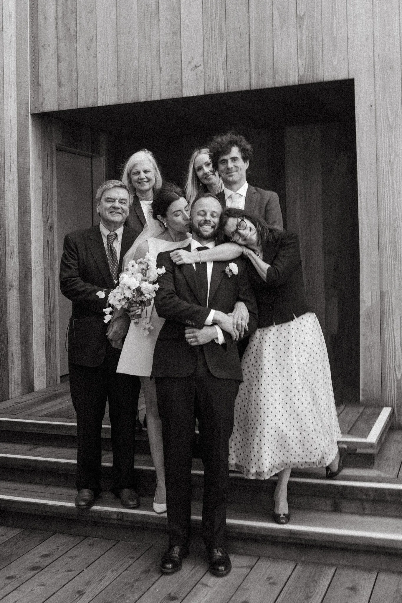 Group of people posing in formal attire on wooden steps, featuring a bride and groom at the center. The image is in black and white.