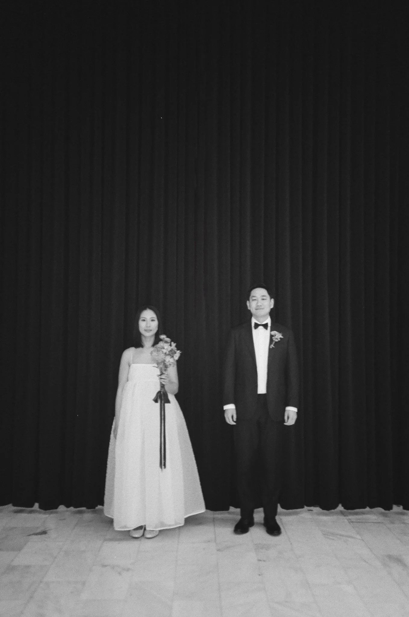 Black and white photo of a bride and groom standing side by side on a stage with a dark curtain background. The bride is holding a bouquet of flowers and wears a strapless wedding gown. The groom is dressed in a tuxedo with a bow tie.
