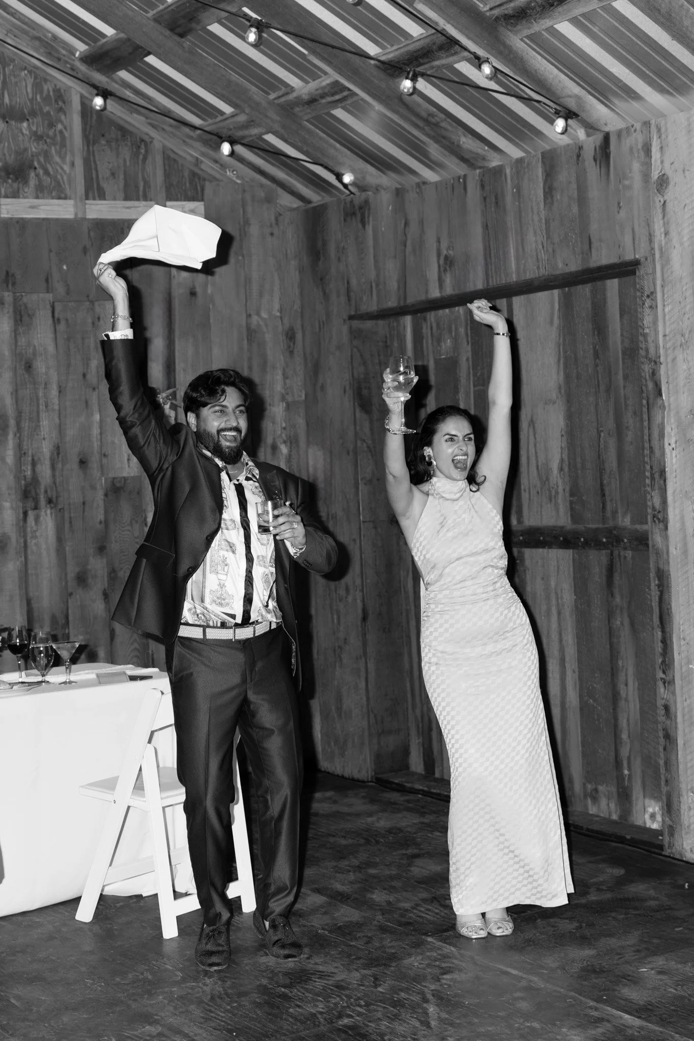 A man in a suit and a woman in a dress celebrate with drinks in a rustic wooden room, smiling and raising their arms.