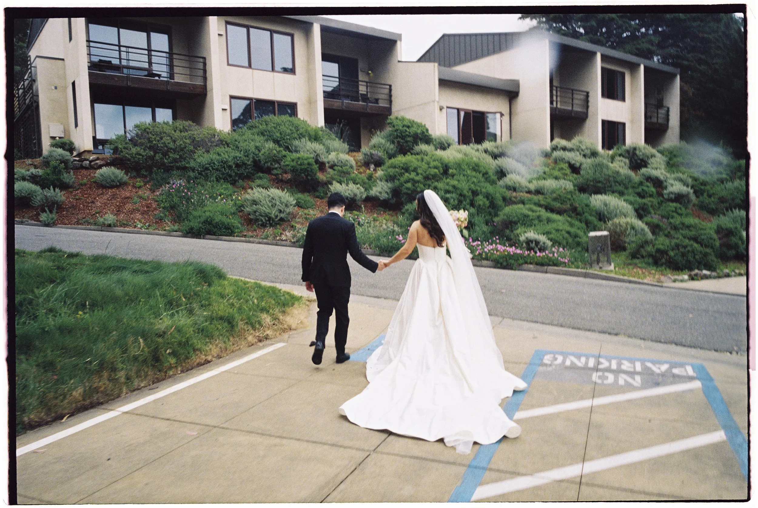 Couple in wedding attire walking hand in hand on a sidewalk near a landscaped apartment complex with greenery and flowering bushes.