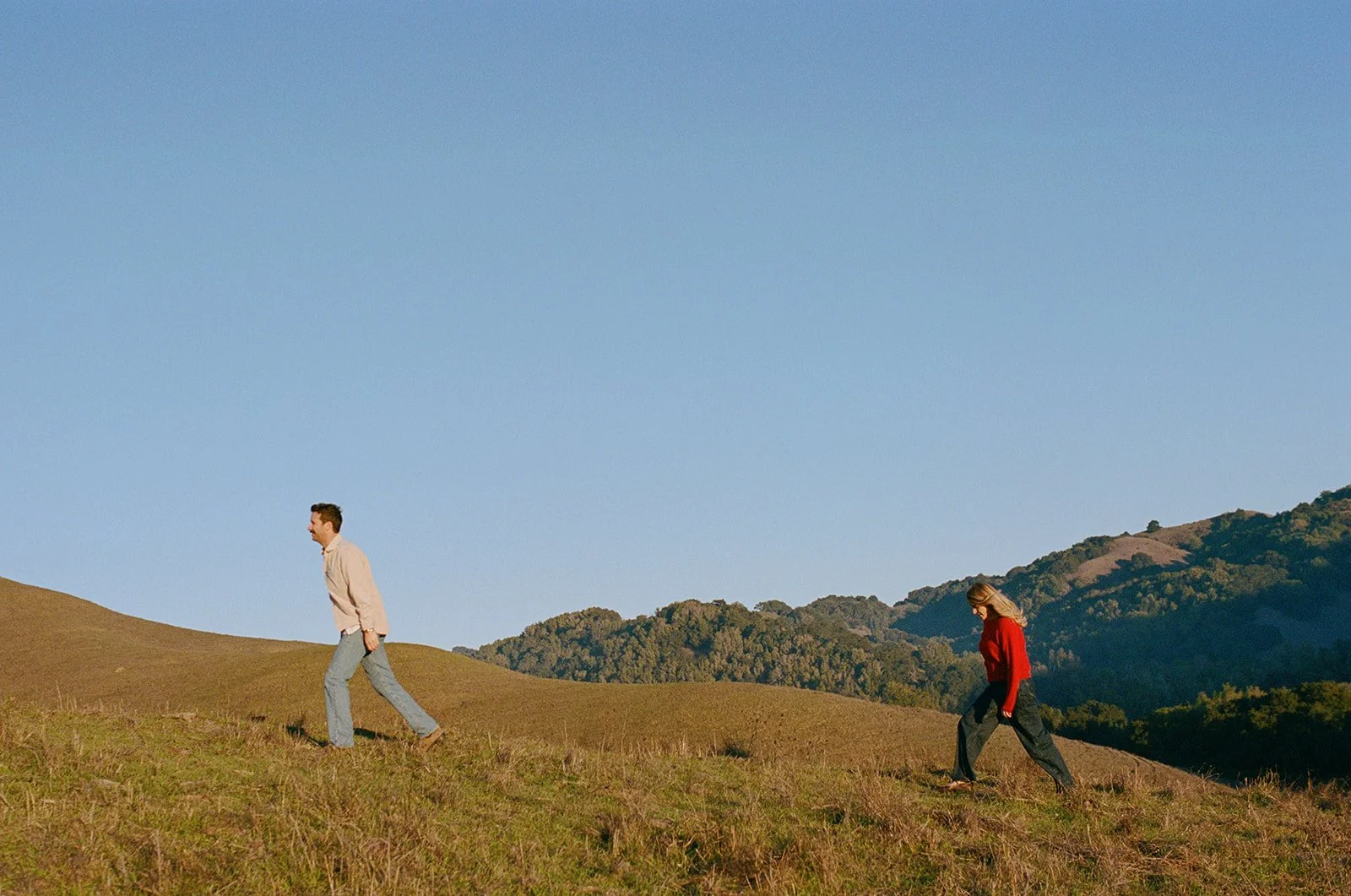 A man and a woman walking uphill on a grassy hillside with mountains in the background under a clear blue sky.