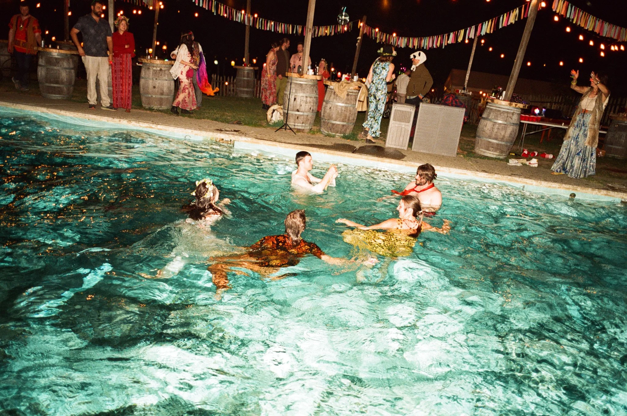 People swimming in a pool at a nighttime party with festive lights and colorful decorations.