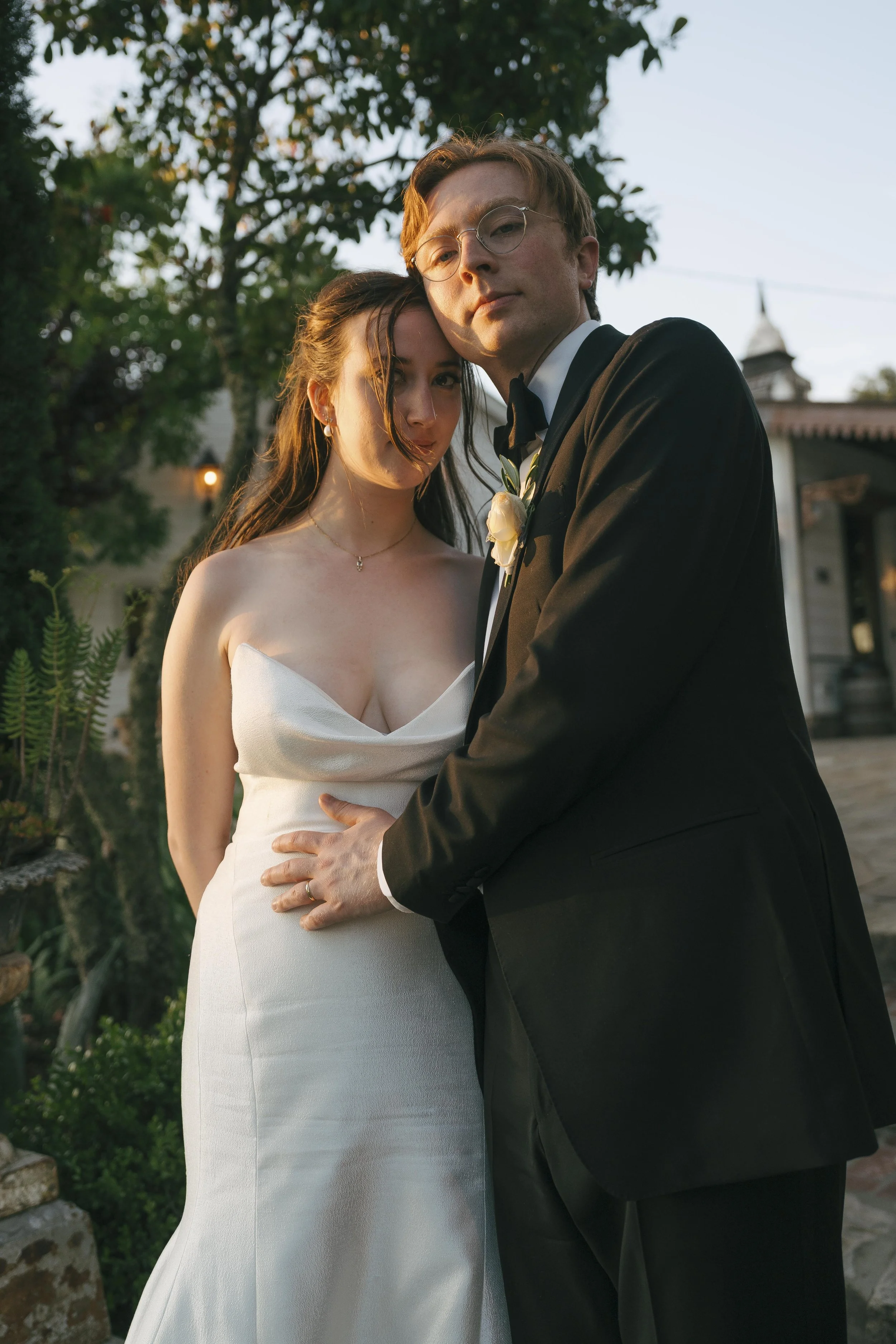 A bride and groom standing close together outdoors, the bride in a white gown and the groom in a tuxedo, facing the camera during sunset.