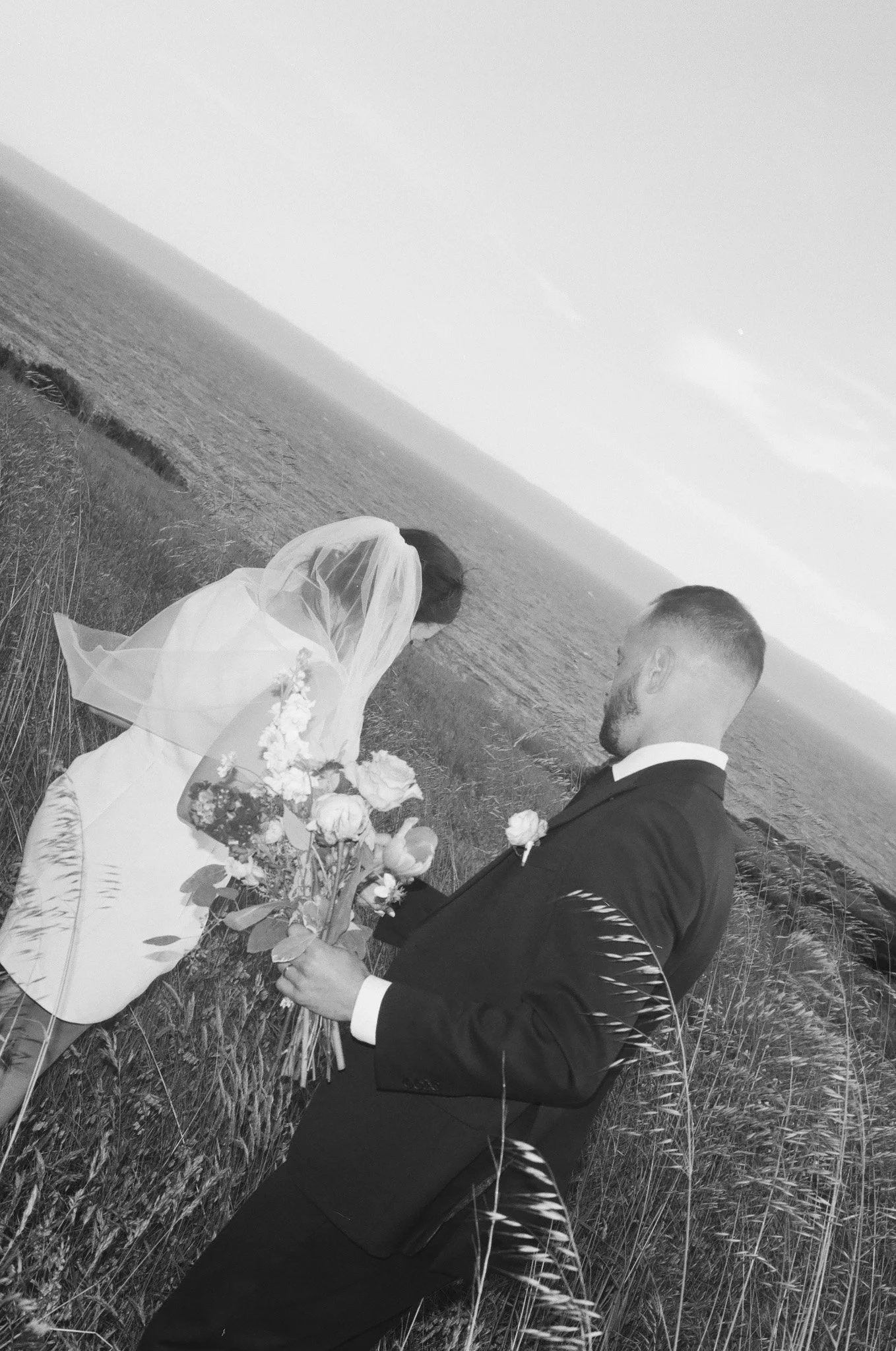 A black-and-white photo of a bride and groom outdoors by the water. The bride is kneeling and holding flowers, wearing a white gown and veil. The groom is standing, holding a bouquet, dressed in a dark suit with a boutonniere, facing the bride at a s