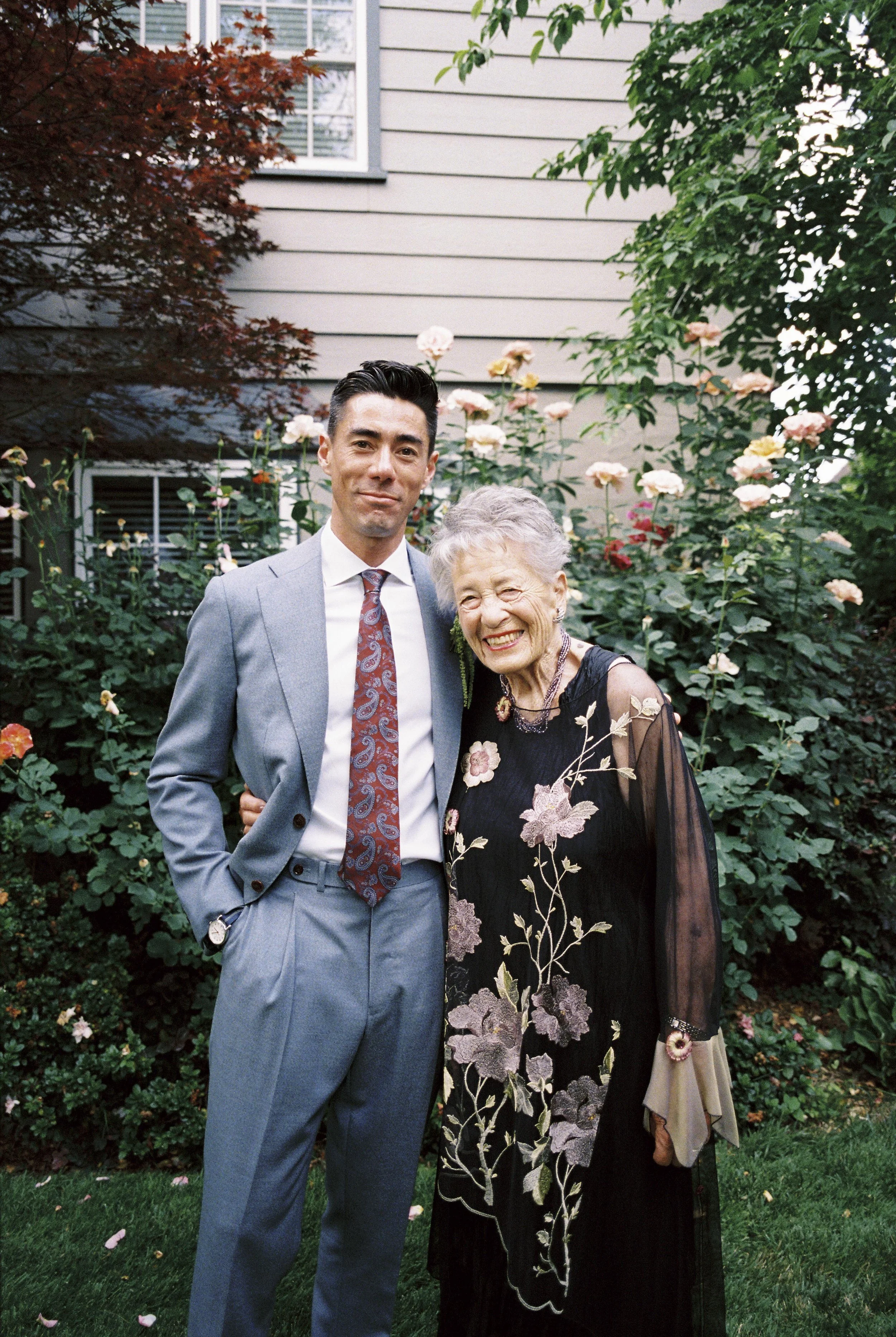 A young man in a light blue suit and a gray patterned tie standing next to an elderly woman in a black floral dress, both smiling in a garden with pink and white flowers and a house in the background.